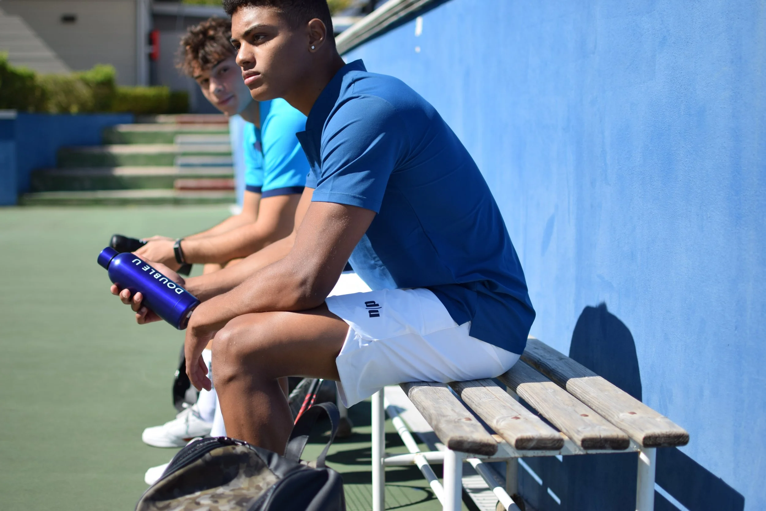 Due giovani uomini seduti su una panchina di legno fuori in un campo da tennis, con magliette blu e pantaloncini bianchi, uno di loro tiene una bottiglia d'acqua blu, un altro ha uno zaino ai piedi.