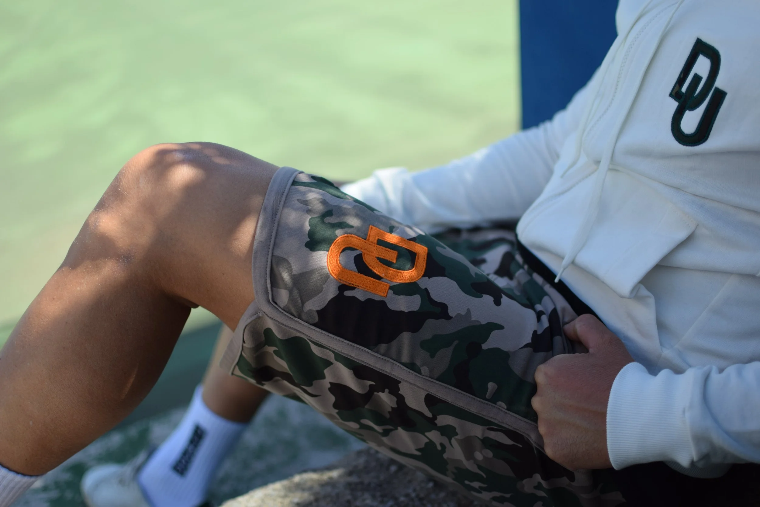 Persona seduta con pantaloni da ginnastica a camouflage, maglietta bianca e calze sportive, con uno sfondo di erba verde.