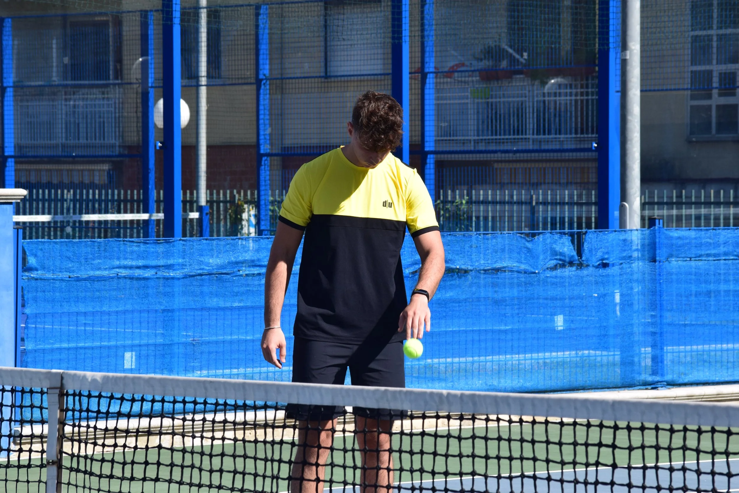 Giocatore di tennis in abito sportivo giallo e nero sulla terra battuta, che tiene in mano una palla da tennis davanti alla rete di un campo.