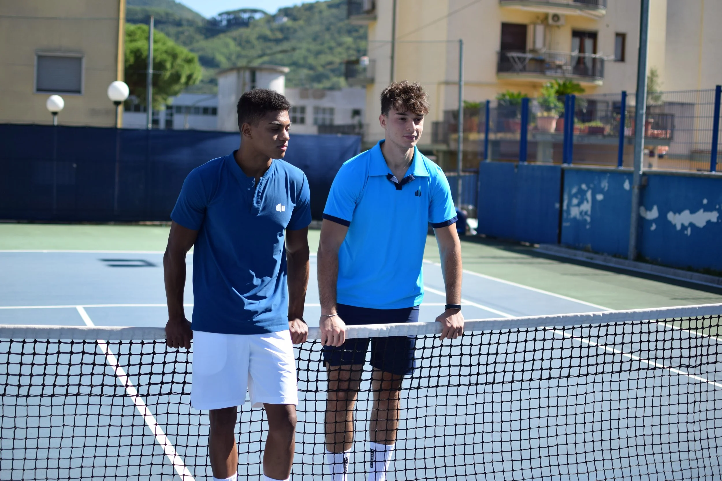 Due giovani uomini si tengono per mano sulla rete di un campo da tennis, pronti a giocare, in un ambiente all'aperto con edifici e colline sullo sfondo.