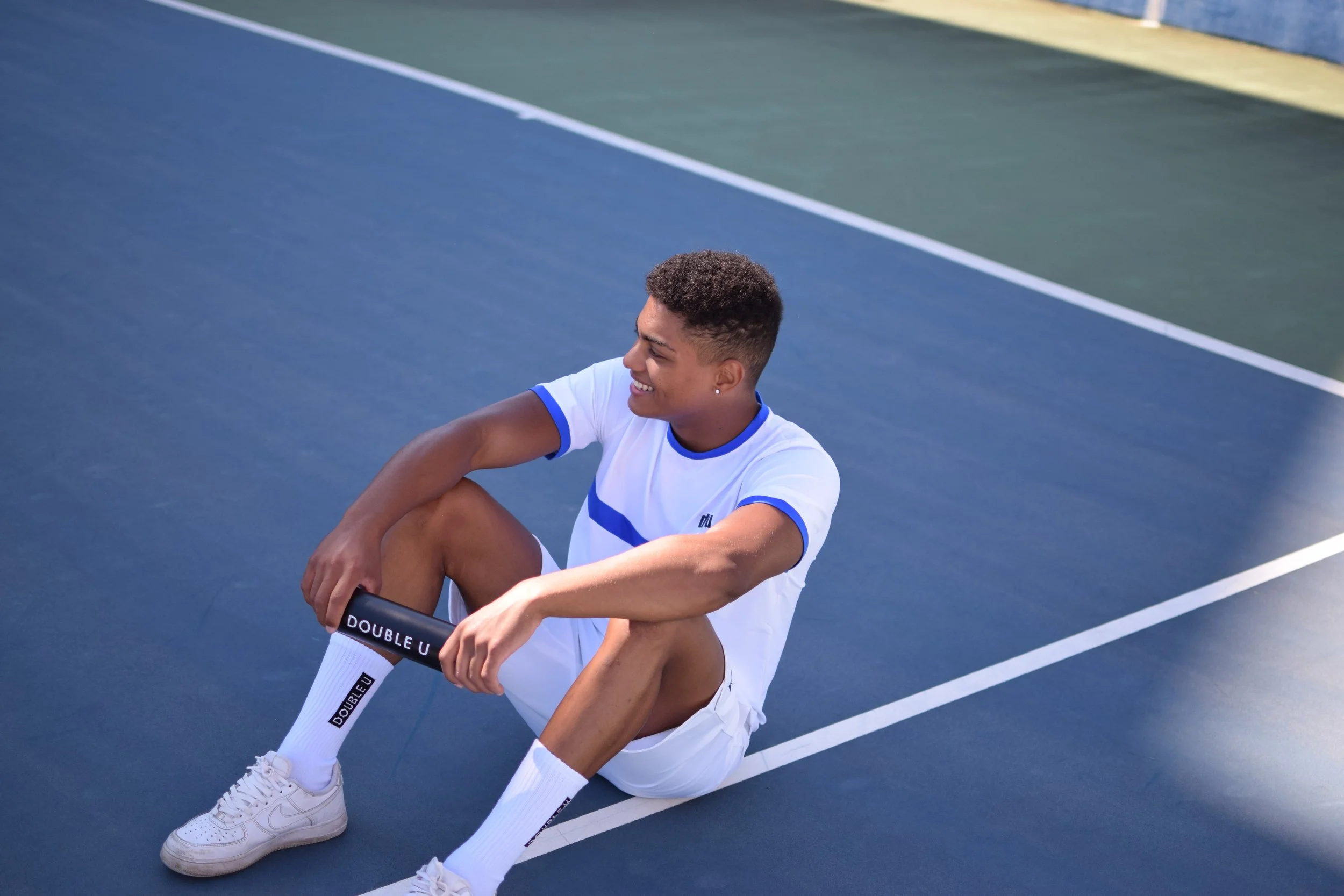 Giovane atleta seduto sul campo da tennis, sorridente, indossa abbigliamento sportivo bianco e calze con scritta 'DOUBLE U', in un campo da tennis all'aperto.
