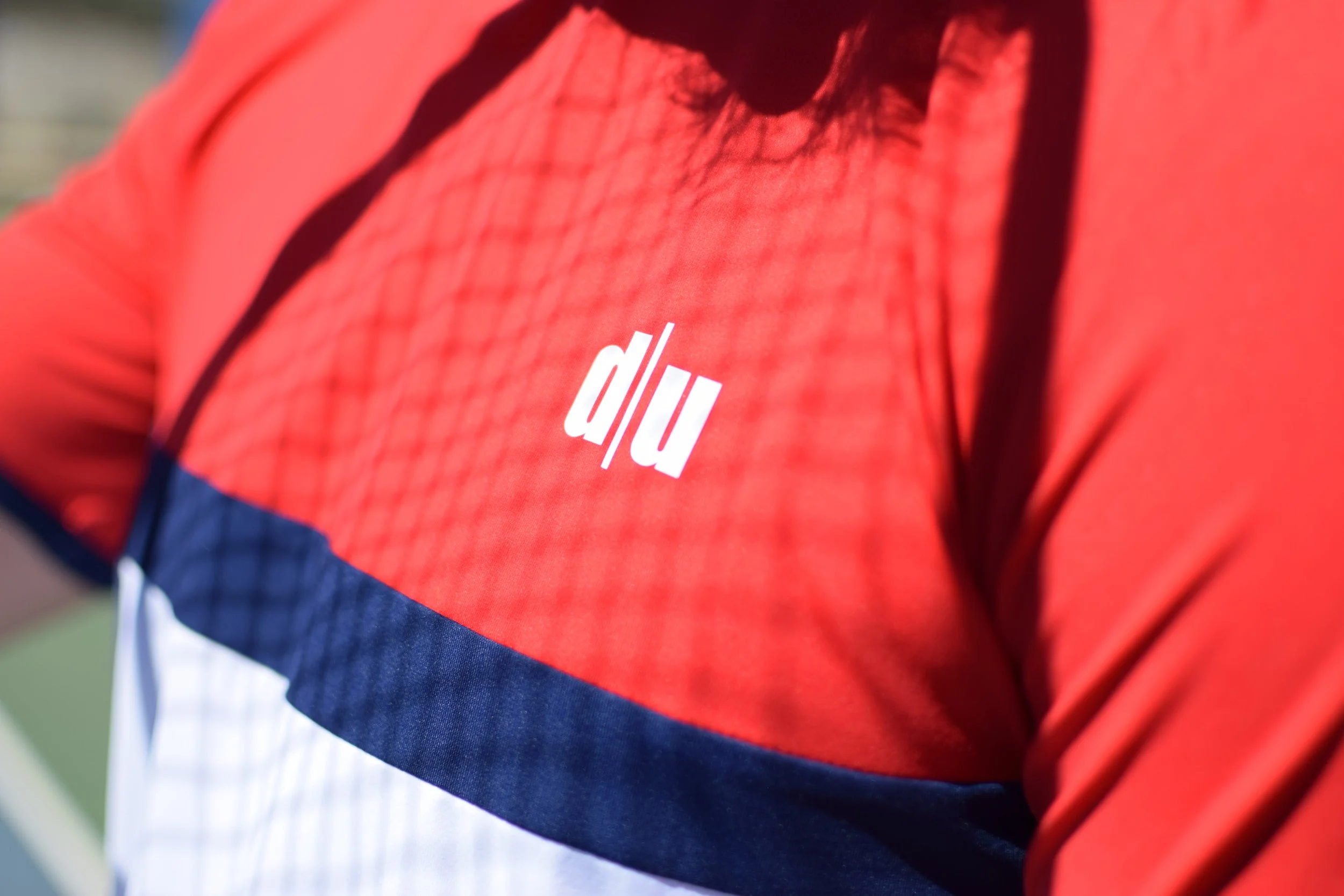 Close-up di una persona che indossa una maglietta rossa con decorazioni nere, blu e bianche, con il logo bianco 'd/l u' sul petto.