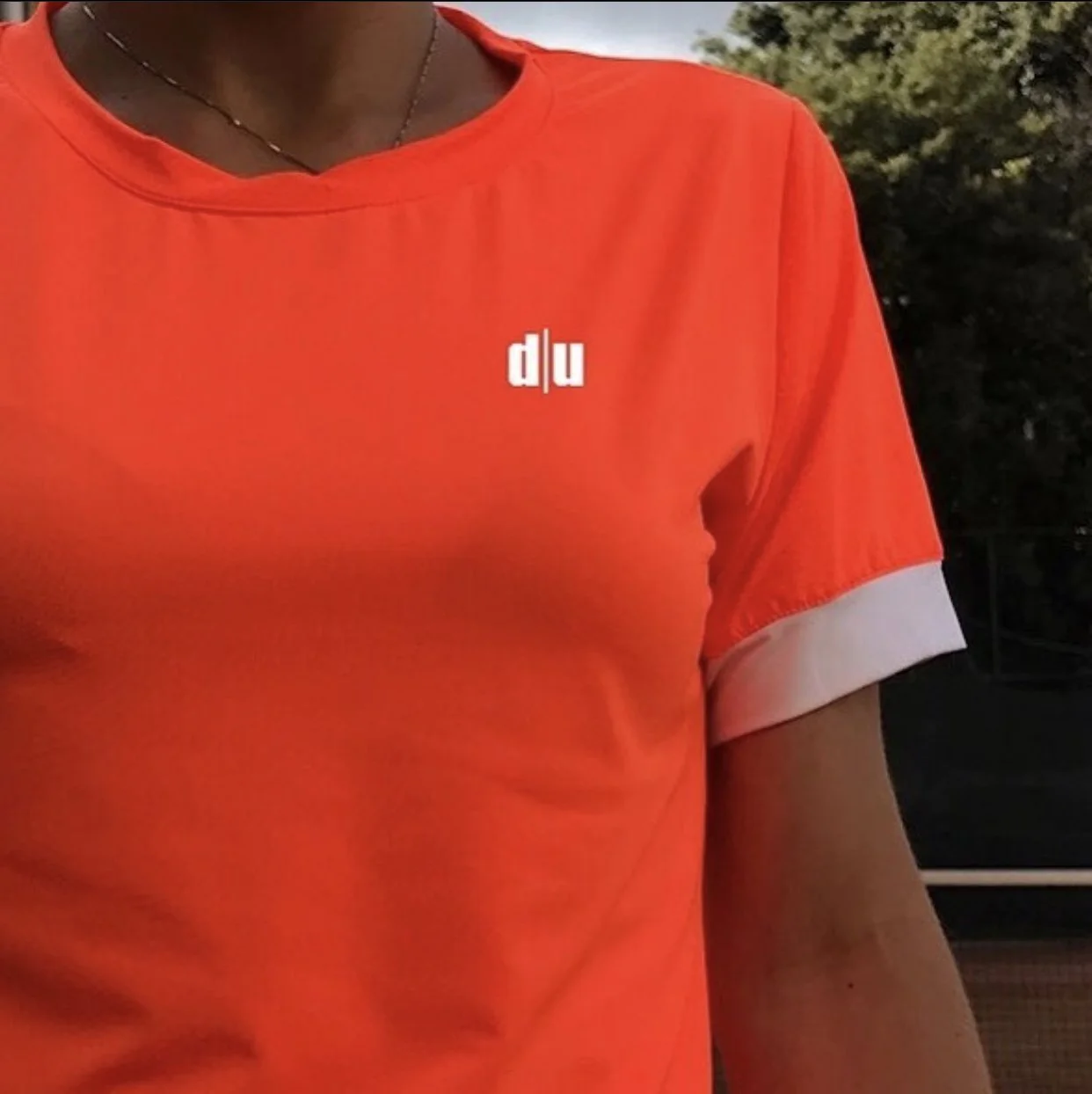 Una persona indossa una maglietta arancione con il logo bianco 'd/u' sul petto, con lo sfondo di natura e cielo.