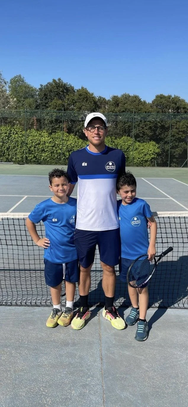 Un uomo e due bambini su un campo da tennis, sorridenti, pronti per giocare, in magliette sportive blu, con rete da tennis davanti e un cielo sereno sullo sfondo.