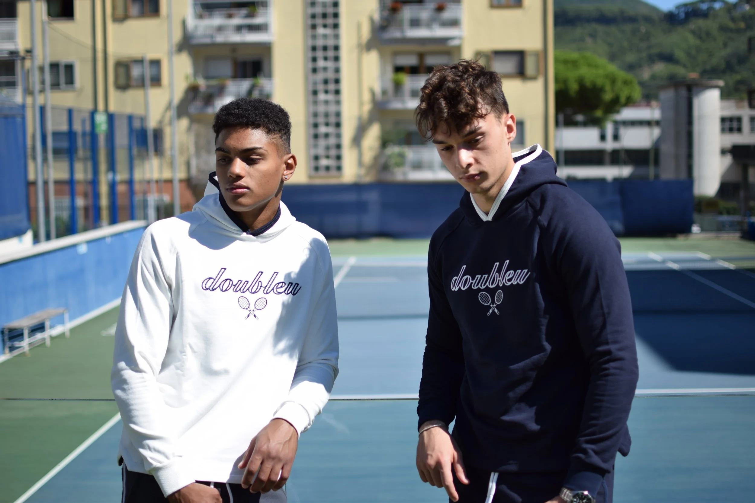 Due giovani uomini sulla pista da tennis, entrambi indossano felpe con il logo 'doubleu' e due racchette incrociate, sullo sfondo ci sono edifici e un cielo sereno.