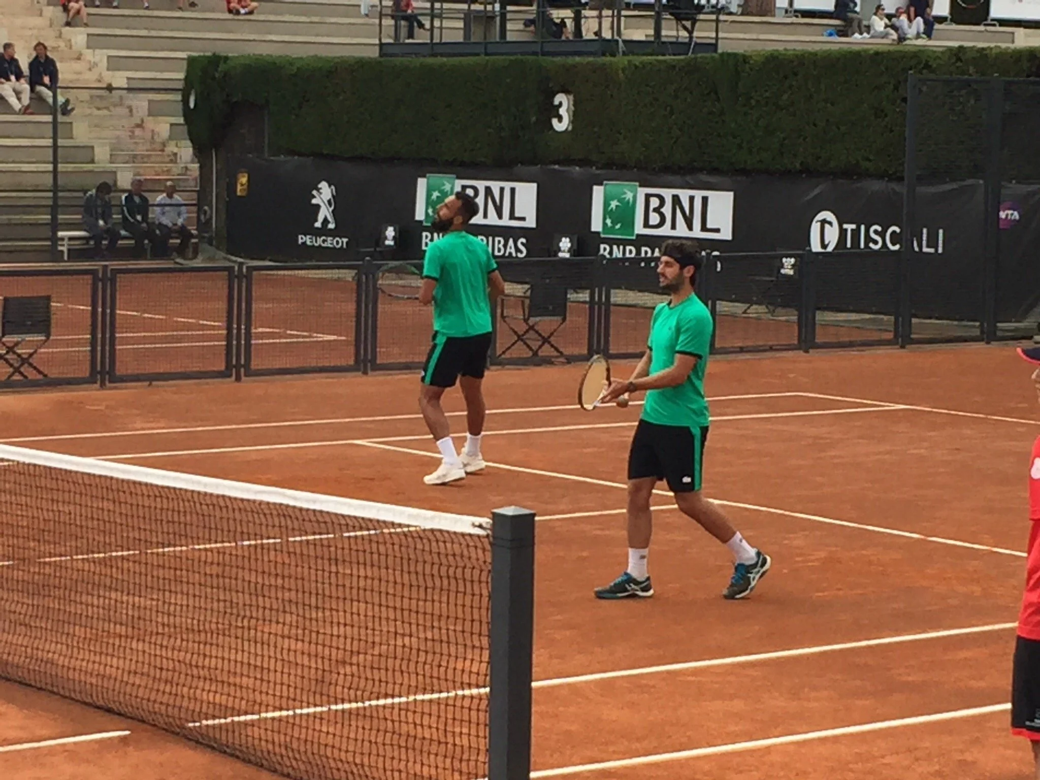 Due uomini in abbigliamento sportivo giocano a tennis su un campo in terra battuta, uno con racchetta in mano.
