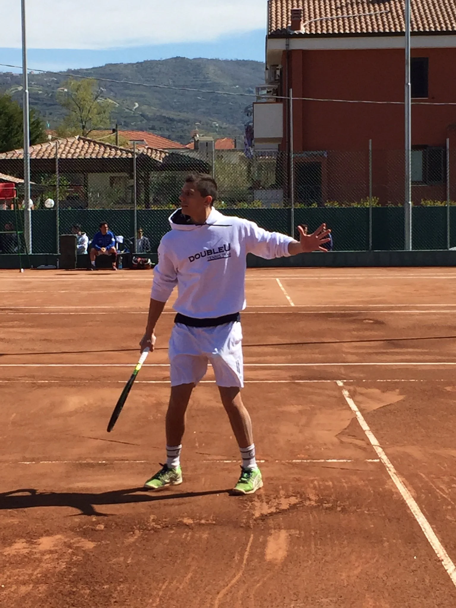 Giovane uomo che gioca a tennis su un campo in terra battuta, con sfondo di case e montagne.