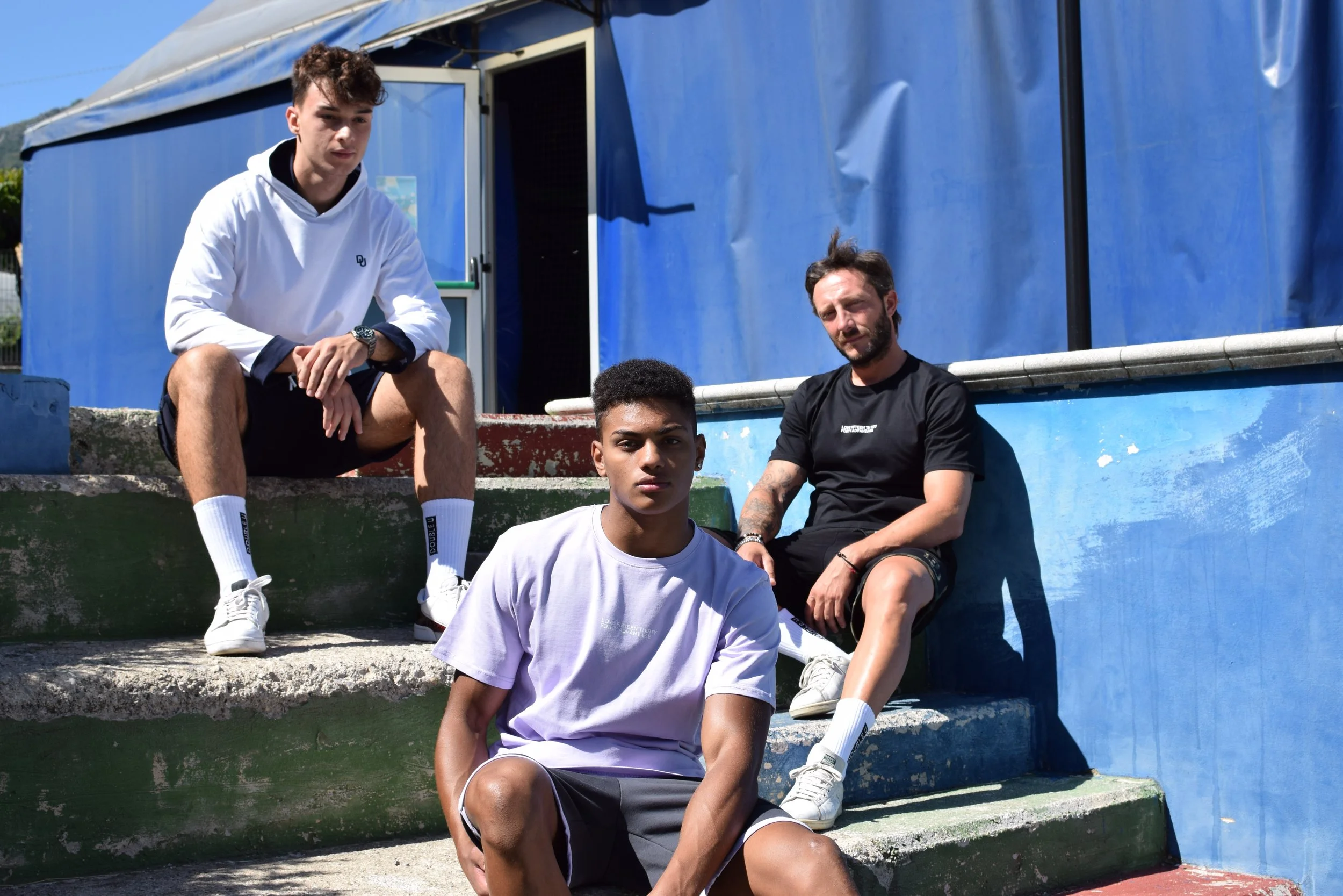 Tre giovani uomini seduti su dei gradini di cemento, all'aperto, con uno sfondo di una struttura in tessuto blu, vestiti in abiti casual, in posizione rilassata.