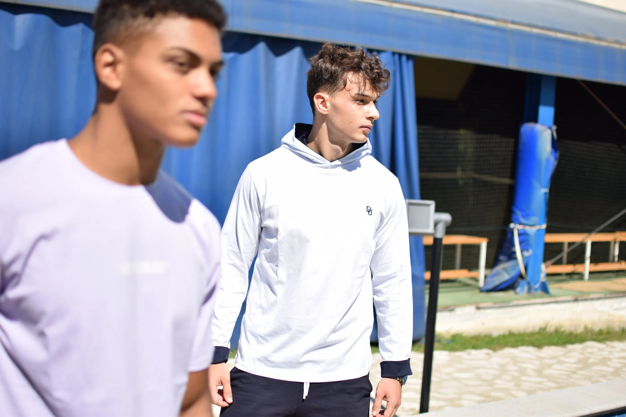 Due giovani uomini con abbigliamento sportivo si trovano all'aperto, uno in primo piano a sinistra leggermente sfocato e l'altro in secondo piano, entrambi con cappotti sportivi e cappucci. Sullo sfondo ci sono tende blu e strutture di allenamento.