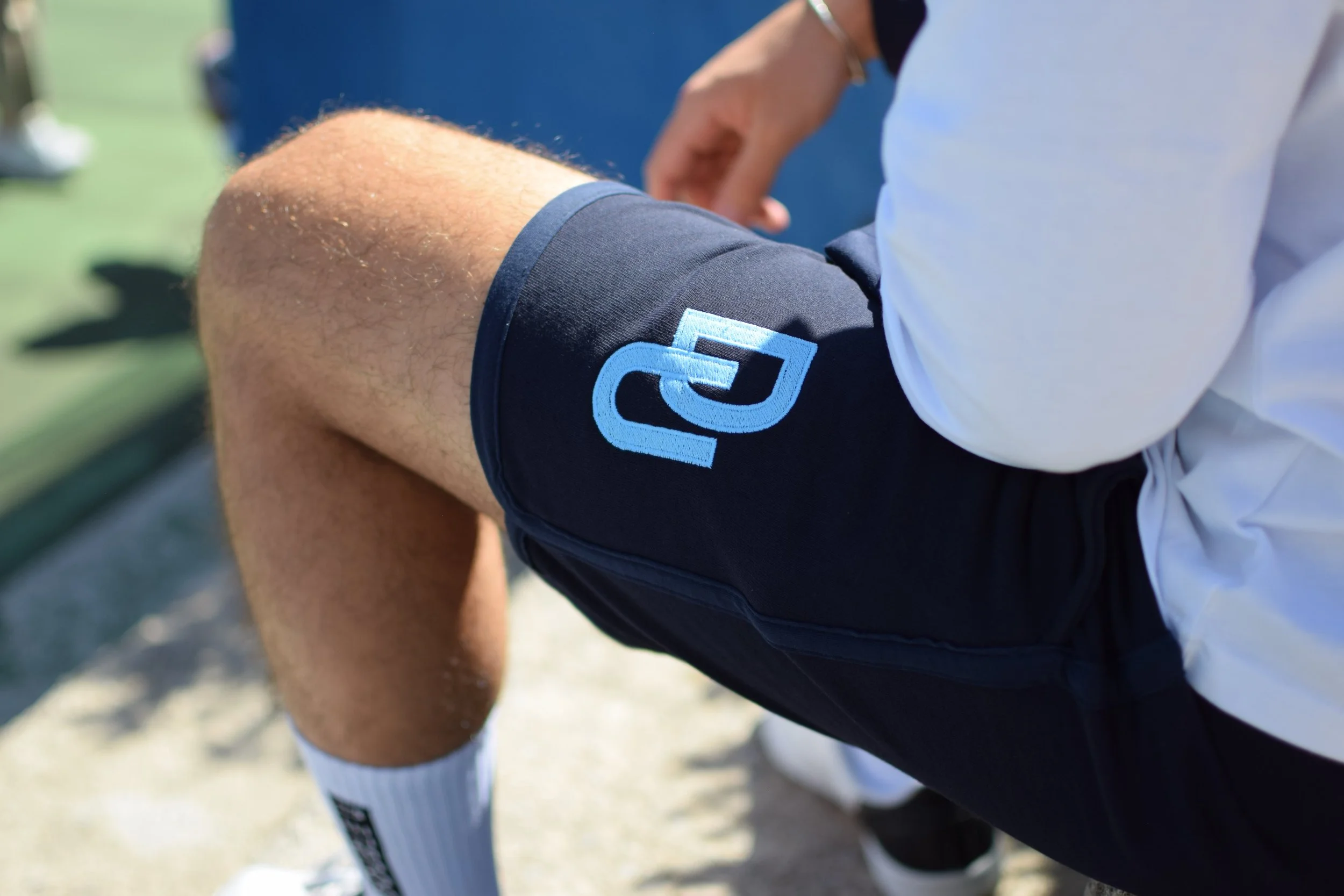 Giocatore di tennis indossa pantaloncini con logo blu sulla coscia, seduto durante una pausa.