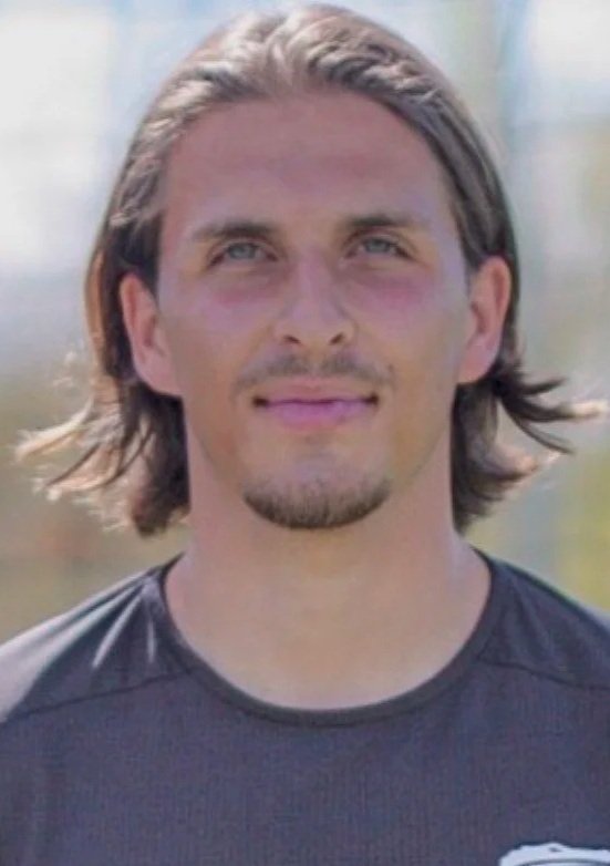 Homme avec des cheveux longs, barbe mince, portant un t-shirt noir, en extérieur, fond flou.