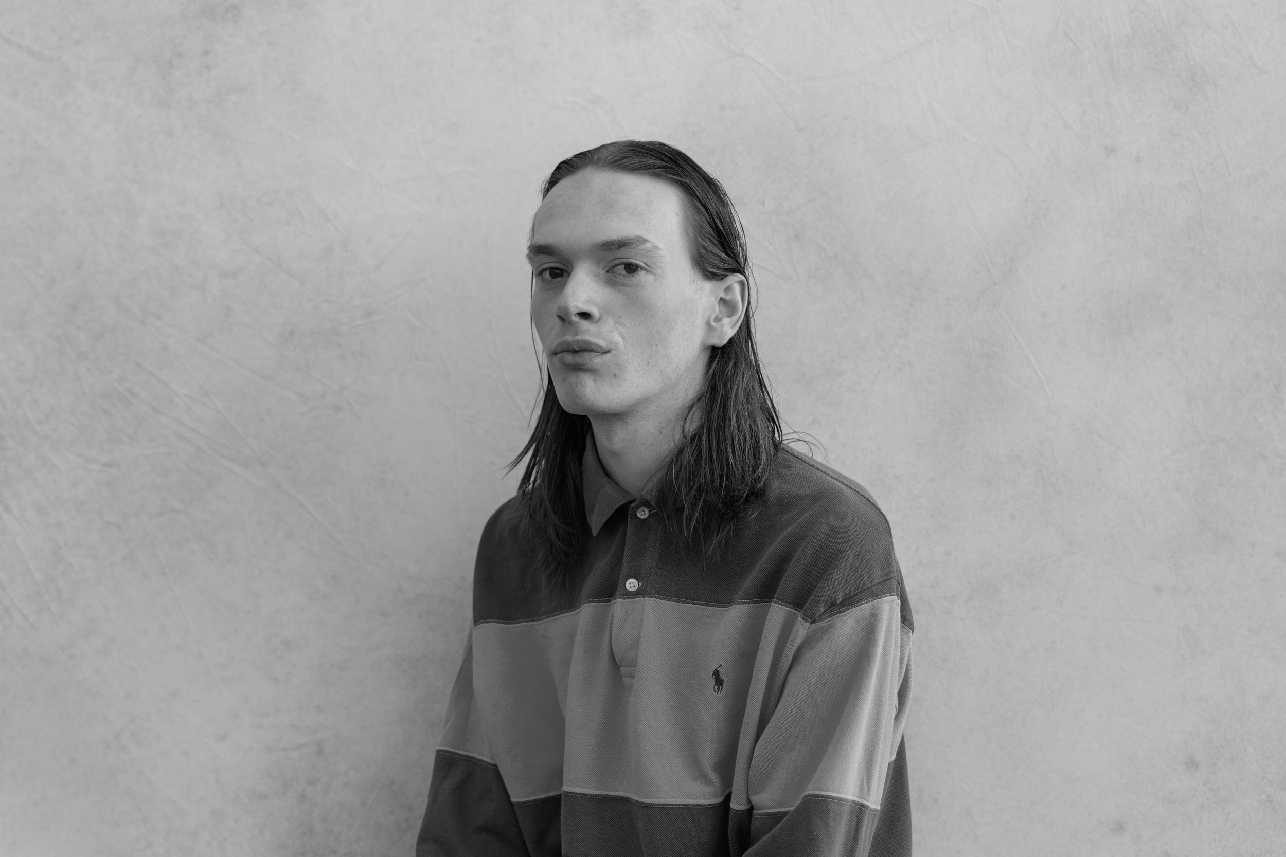 Black and white portrait of a young man with long hair, wearing a polo shirt, standing against a plain wall.