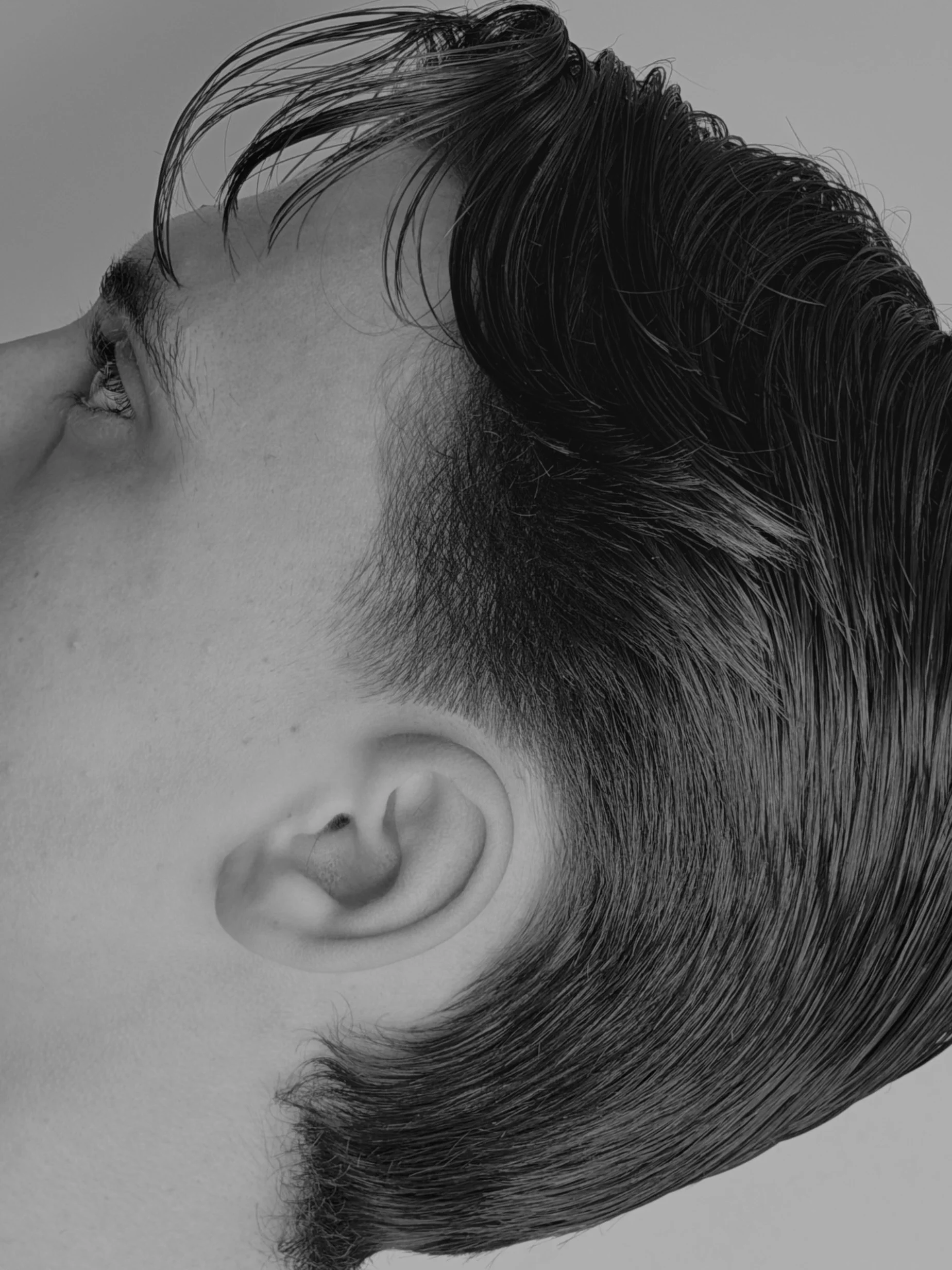 Close-up black and white photo of a person with short, dark hair and a visible ear, lying on a flat surface.