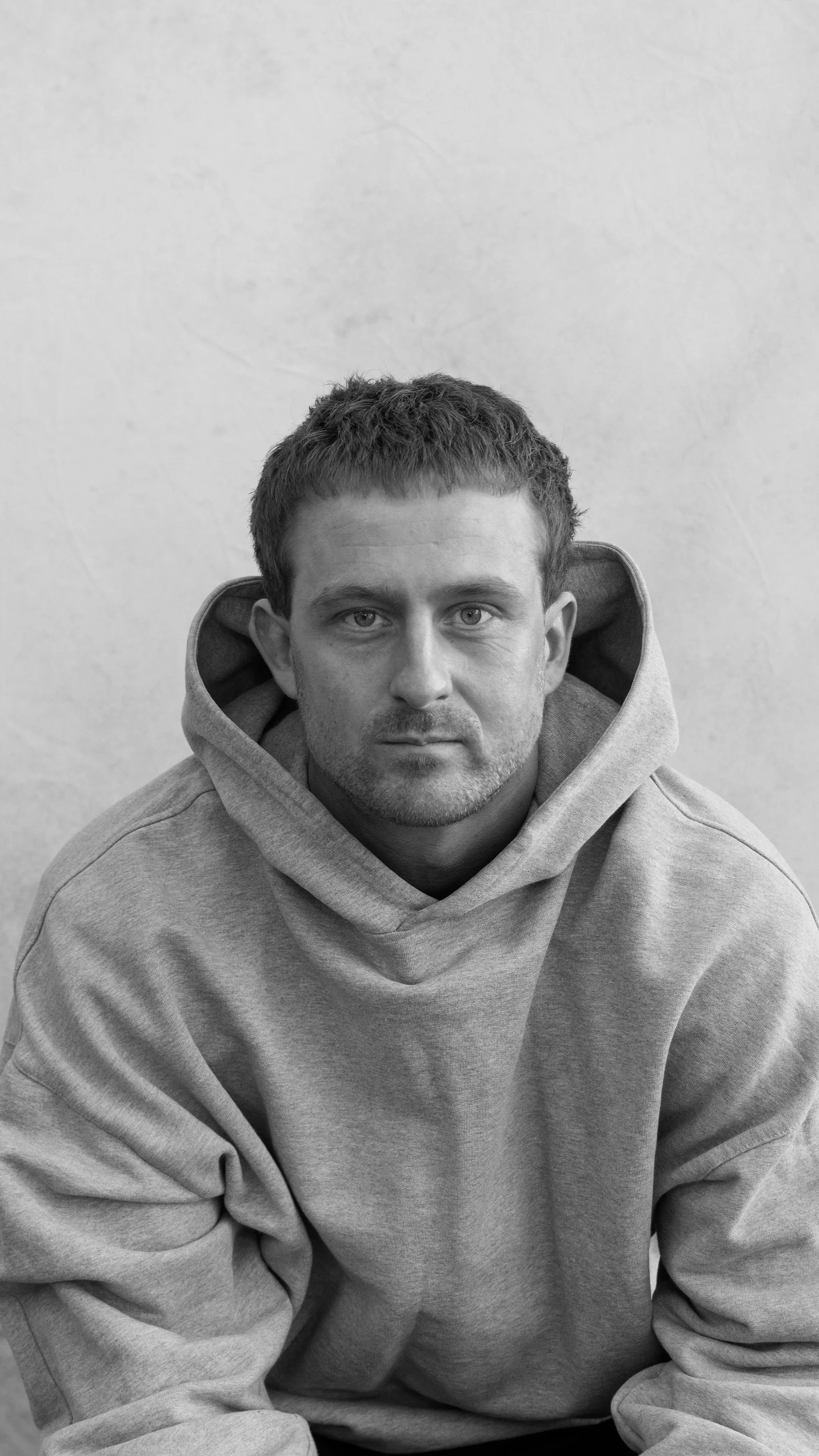 A black-and-white photo of a young man with short, curly hair, wearing a hoodie, sitting against a plain wall, looking directly at the camera.