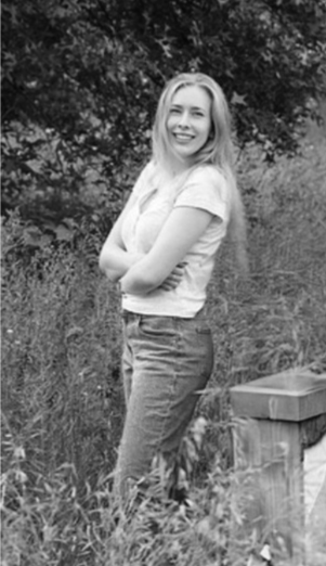 A woman with long hair smiling outdoors, standing with her arms crossed near a wooden fence, in a natural setting with trees and grass.