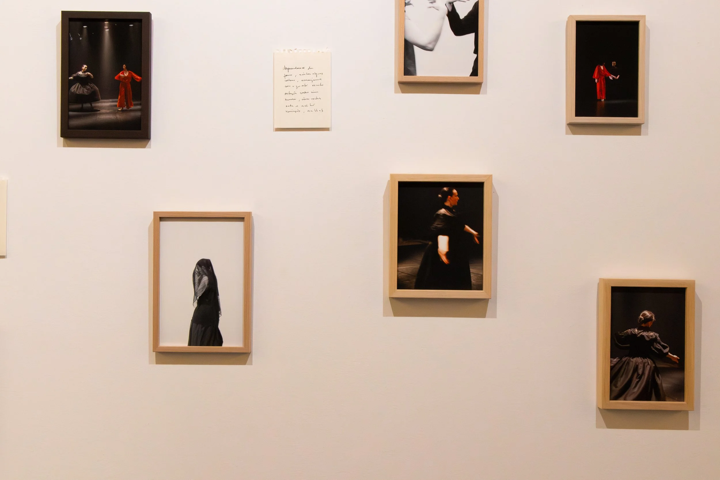 Gallery wall with framed photos of dancers in black costumes, some in motion, against dark backgrounds, with a few uncased images, including a simple handwritten note on white paper.