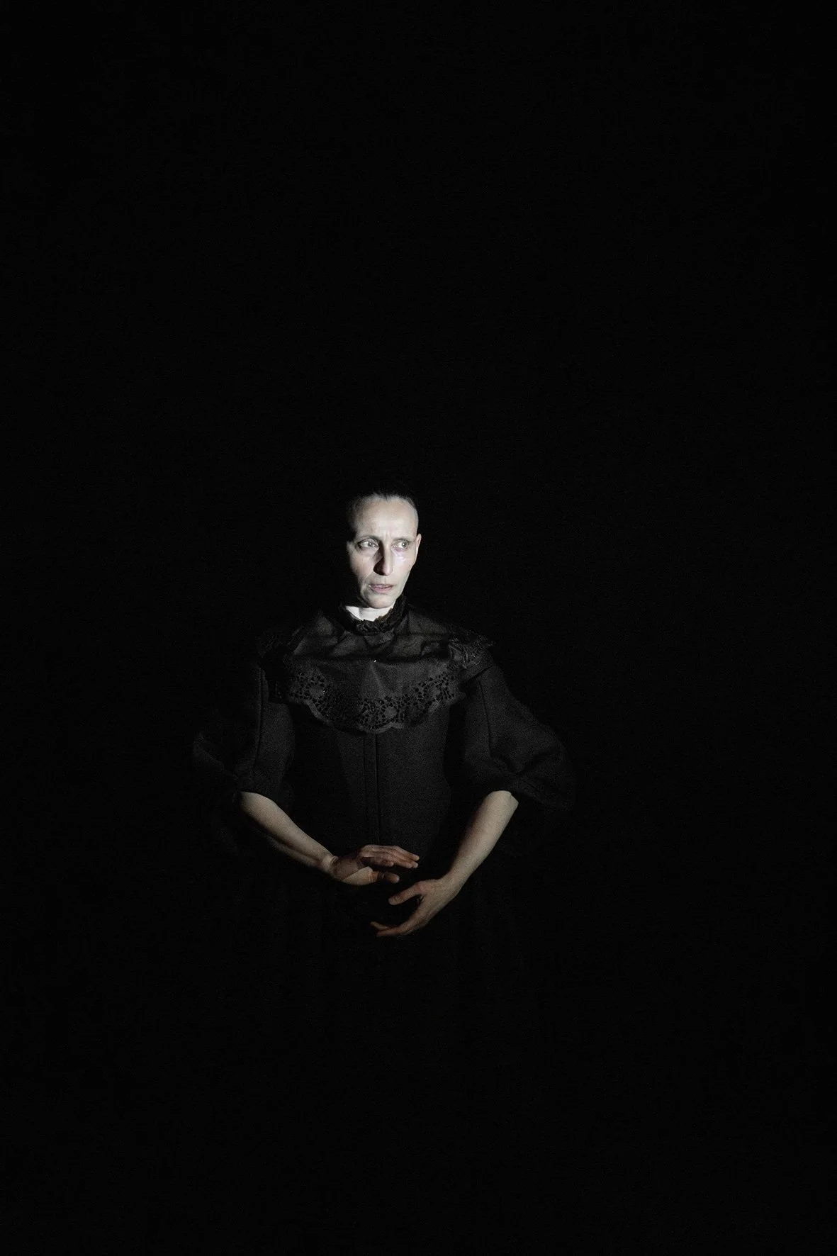 A woman dressed in black with lace details, standing in a dark environment with a focused expression.