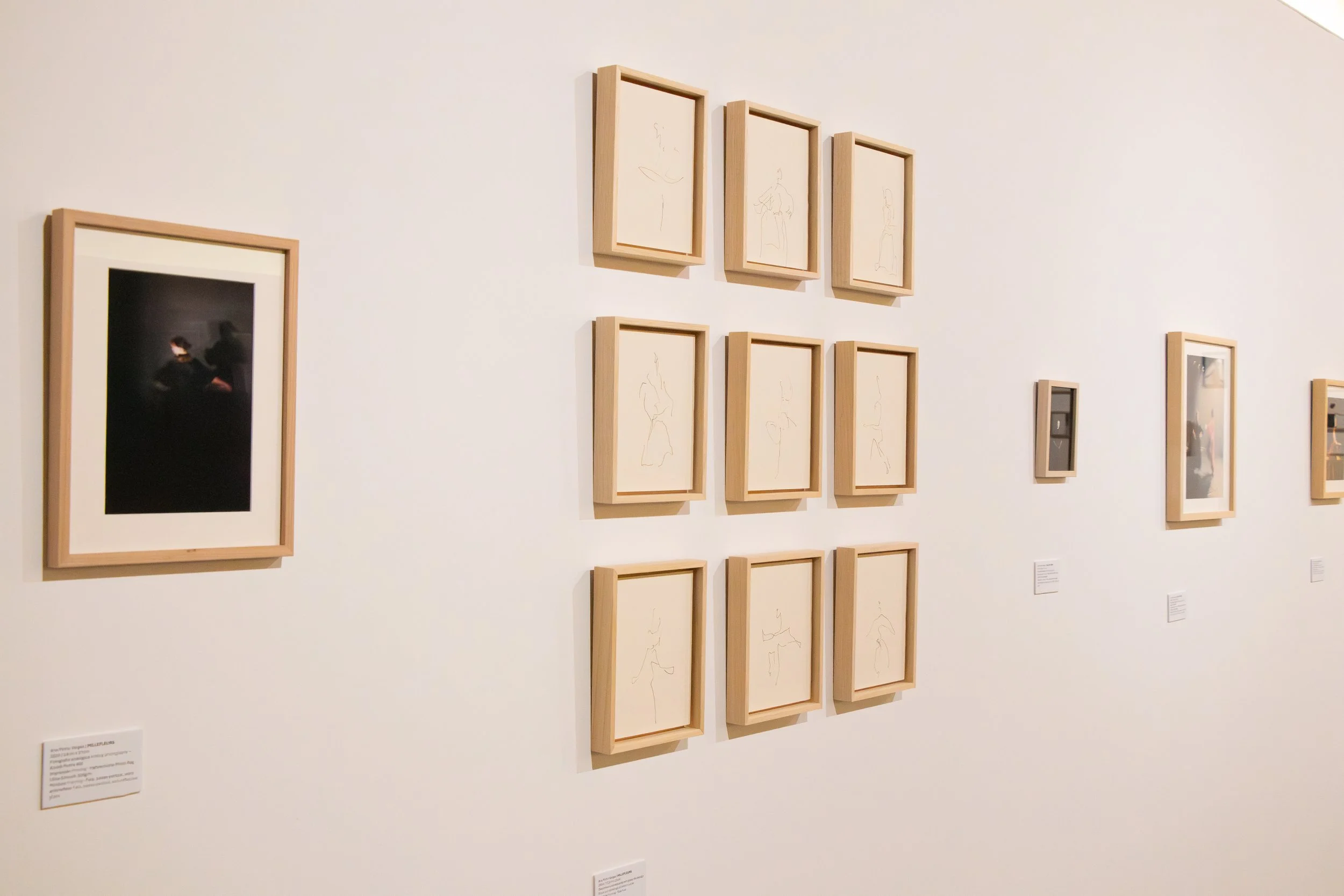 Gallery wall with framed artworks, including a large abstract photograph on the left and smaller sketches of human figures arranged in a grid pattern.