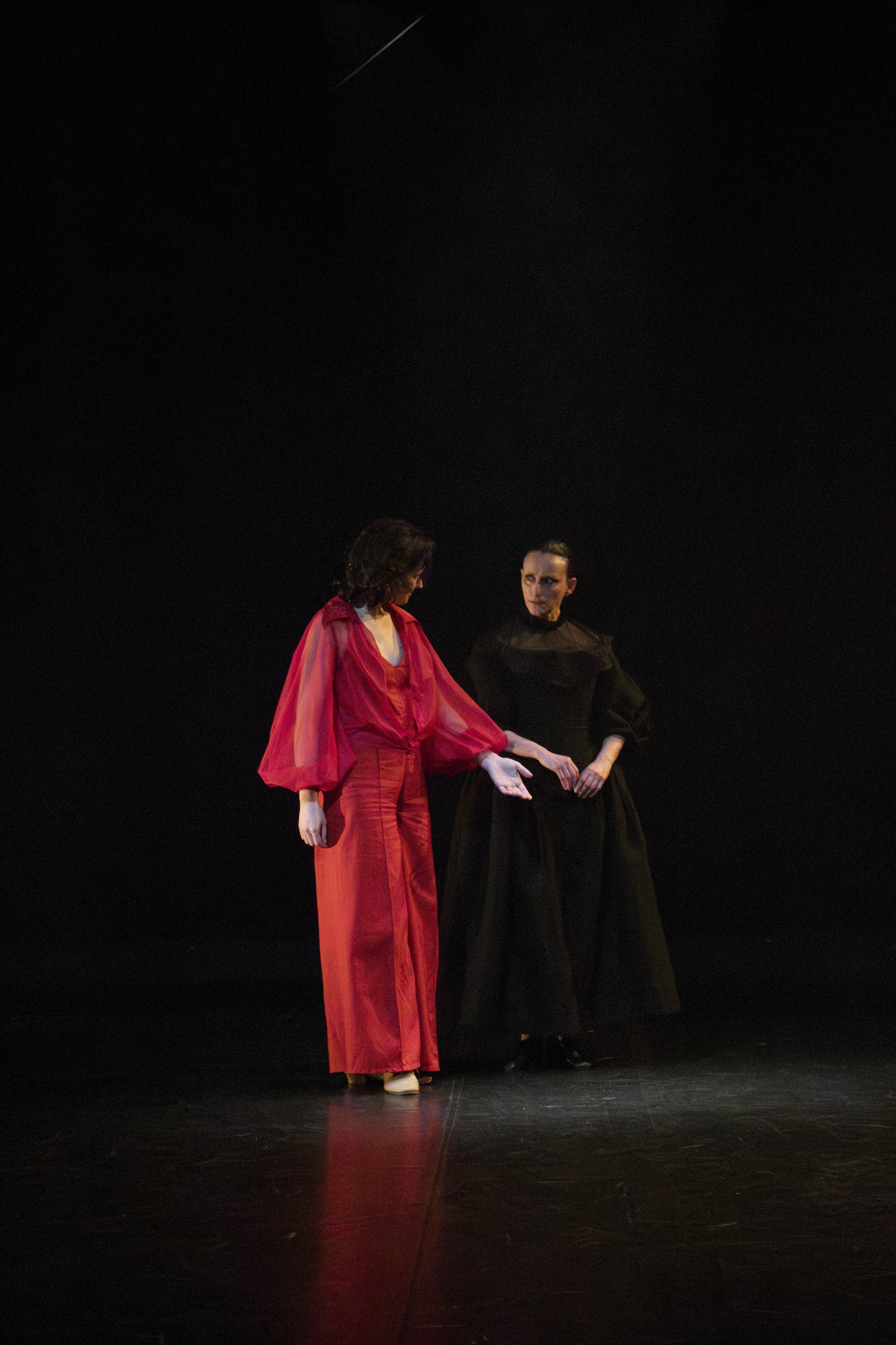 Two women on stage, one in a red dress and the other in a black dress, engaged in a dramatic performance with a dark background.