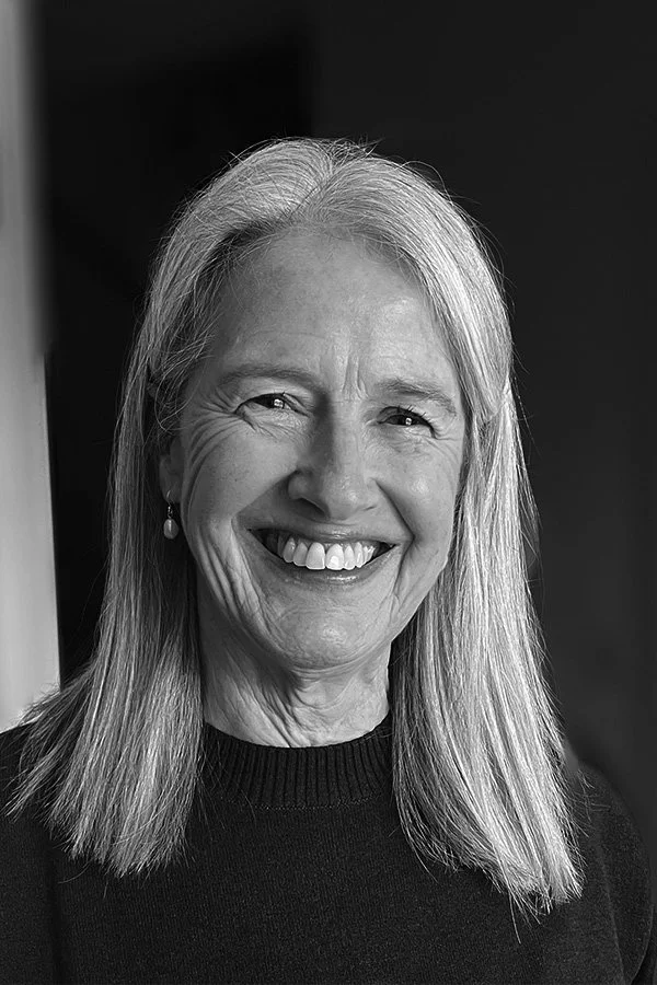 An older woman smiling, wearing a black top with a decorative neckline, standing against a plain wall.