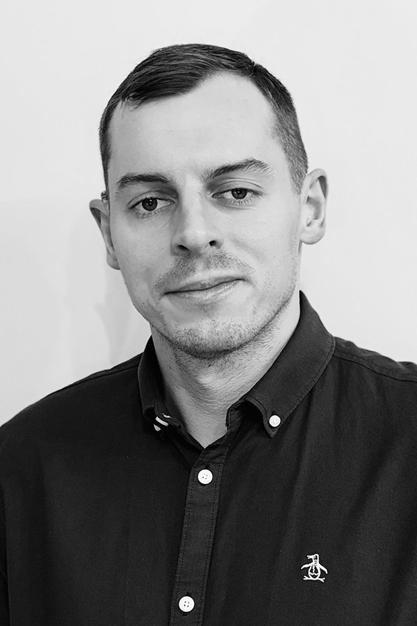 Black and white photo of a young man with short hair, wearing a black crew neck shirt, looking slightly to the side with a neutral expression, standing against a plain light background.