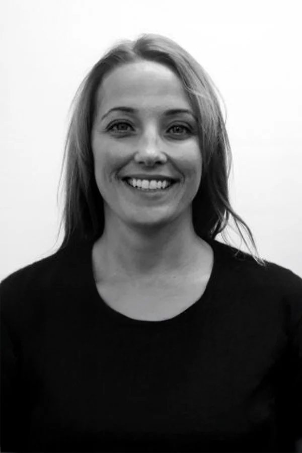 Black and white photo of a smiling woman with shoulder-length hair wearing a dark top, standing against a light background.