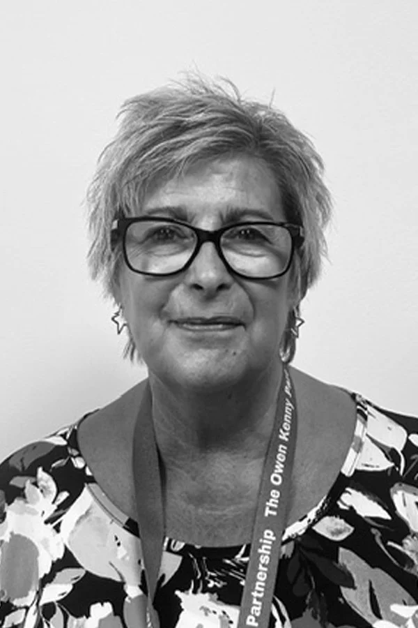 A woman with short, light-colored hair and glasses, wearing a floral-patterned top and a lanyard.