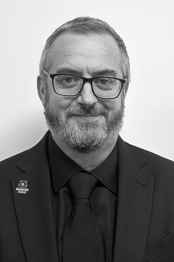 Black and white professional headshot of a man wearing glasses, a dark tie, and a suit jacket, smiling softly at the camera.