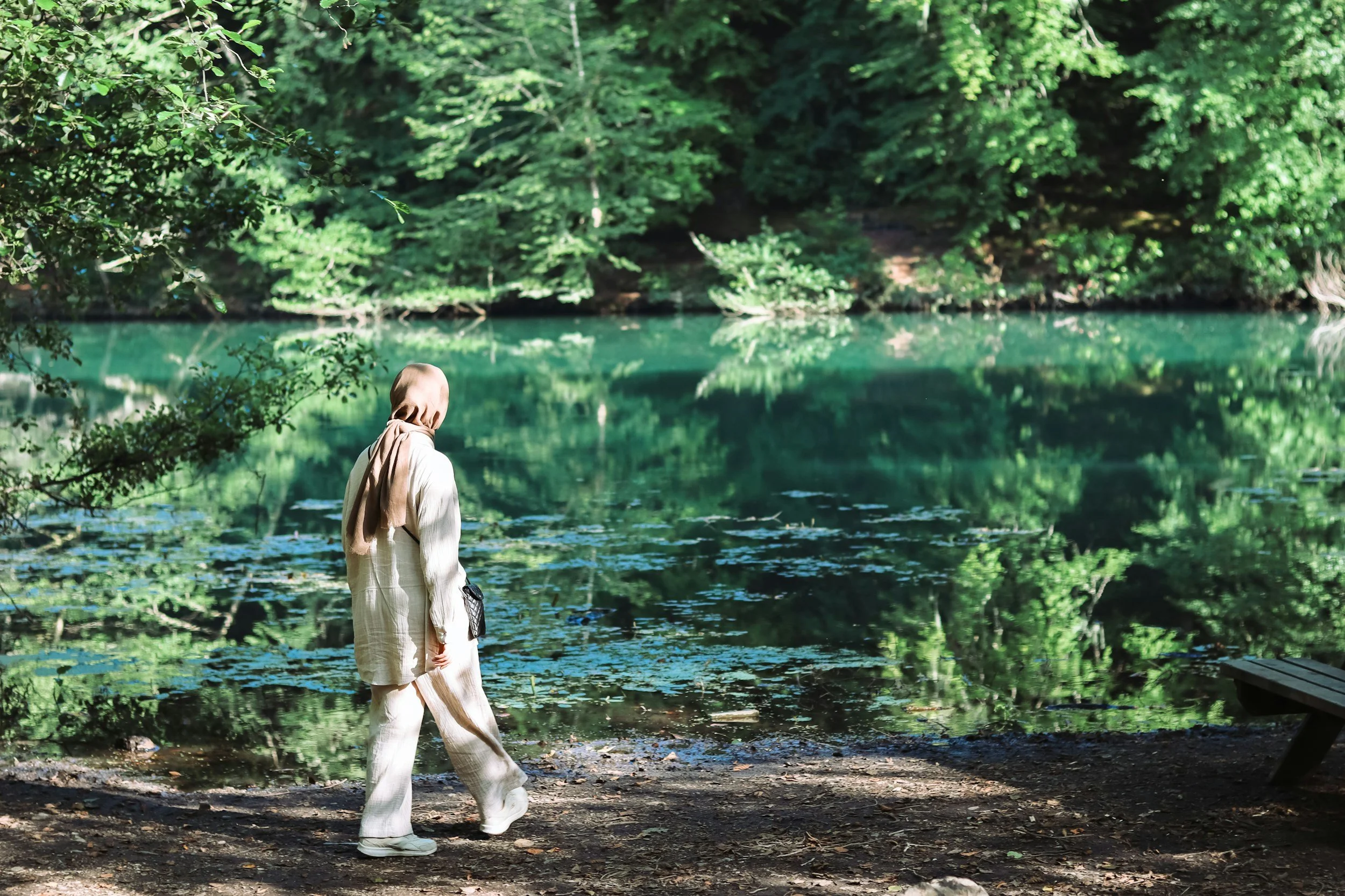 Person in beige and white clothing standing near a body of water in a lush green forested area.