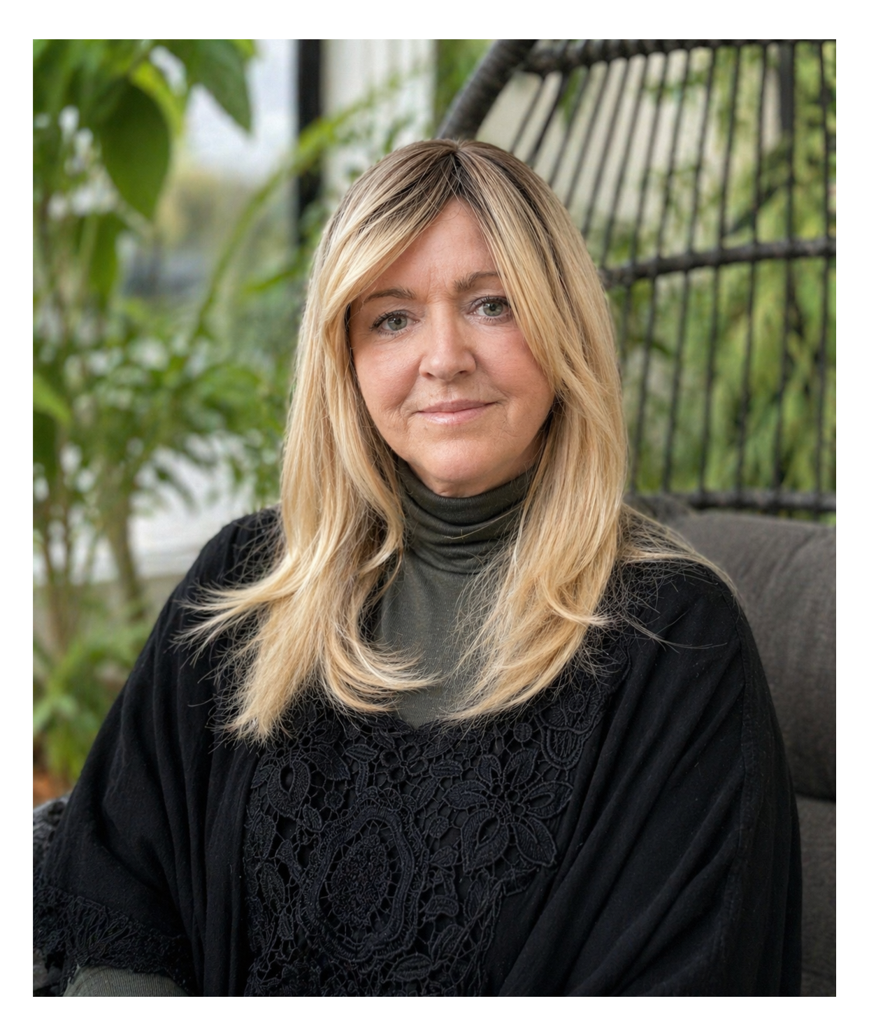 Michelle Courtney, trauma informed psychotherapist sitting on a gray chair in a therapy room with green plants and a forest scenery in the background, wearing a black lace top and a dark turtleneck under it.