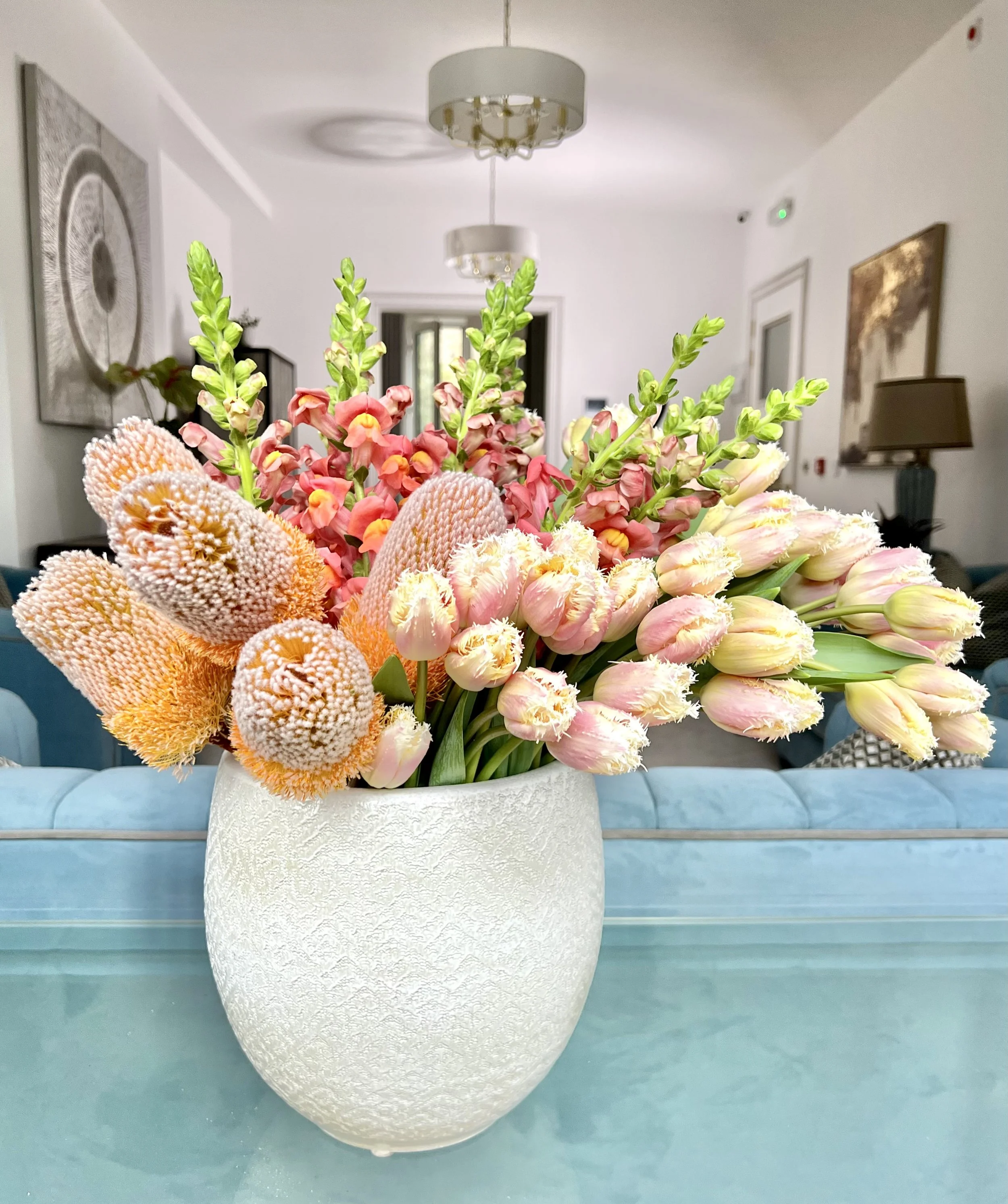 A white vase filled with various flowers, including pink tulips, orange proteas, and green snapdragons, placed on a glass table in a modern living room.