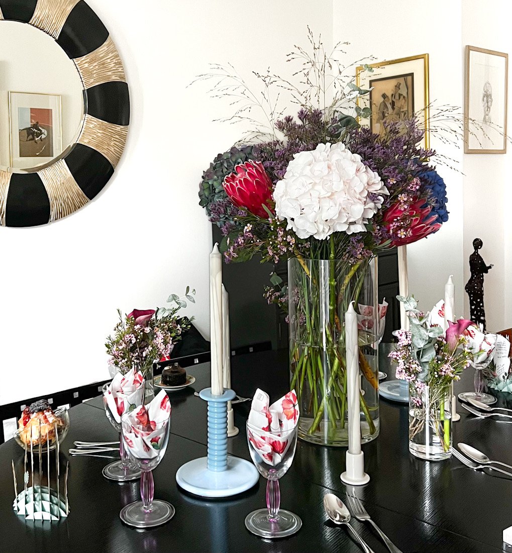 Elegant dining table with large floral centerpiece, candles, decorative glasses with floral napkins, and modern decor.