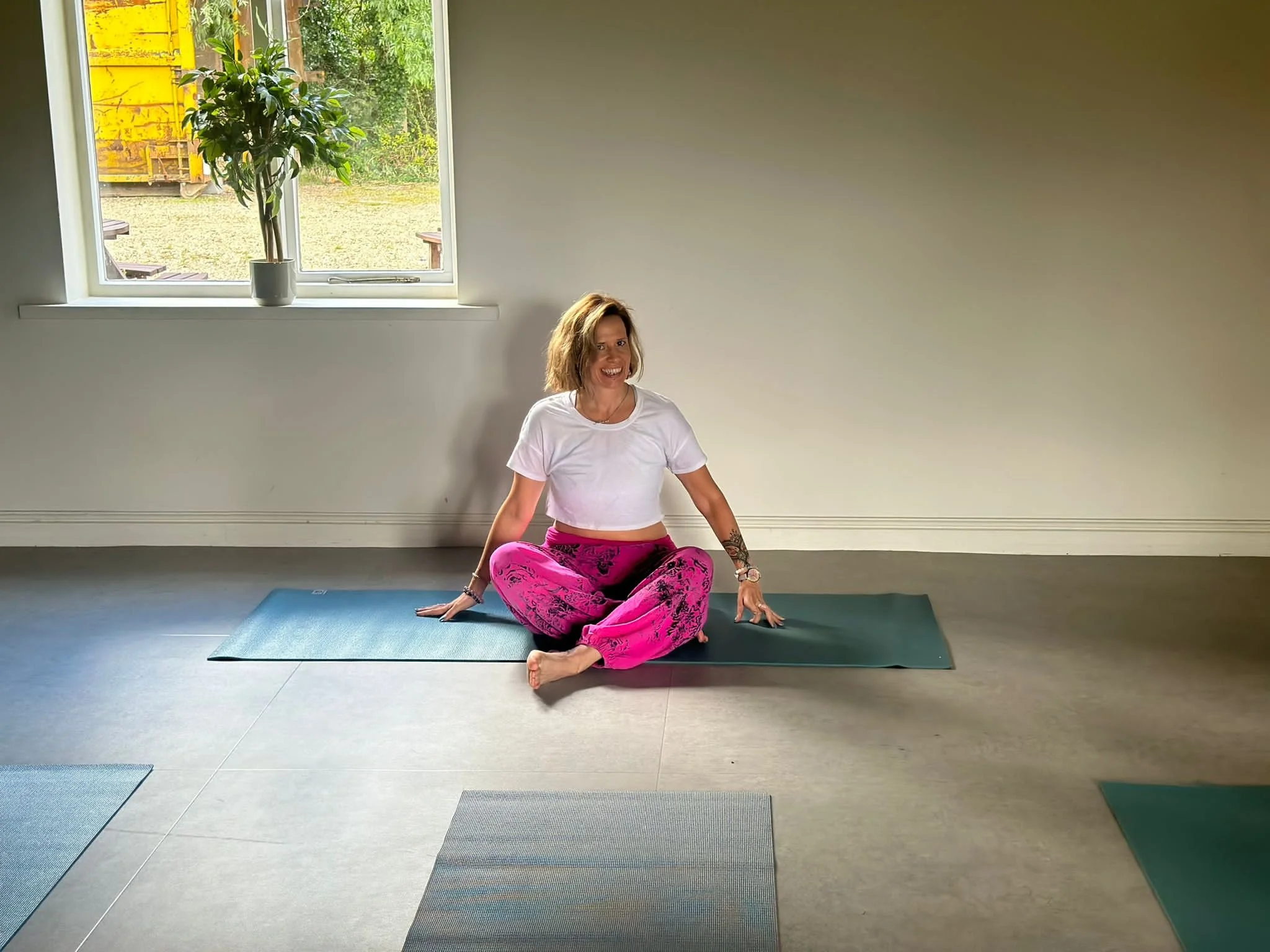 Image of yoga teacher, sitting on floor on yoga mat in cross legged position wearing pink trousers and white top