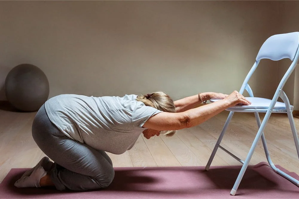 A person kneeling on a yoga mat stretches their arms forward onto a folding chair, lowering their head and chest in a gentle supported stretch.