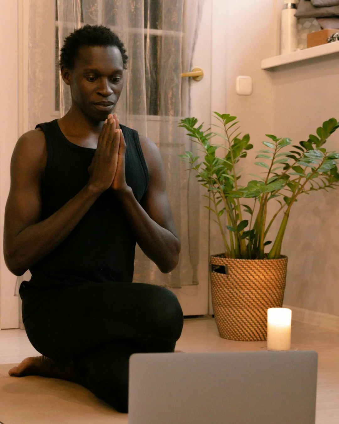 A person doing yoga at home on their laptop with a candle and plant beside them looking focused and calm