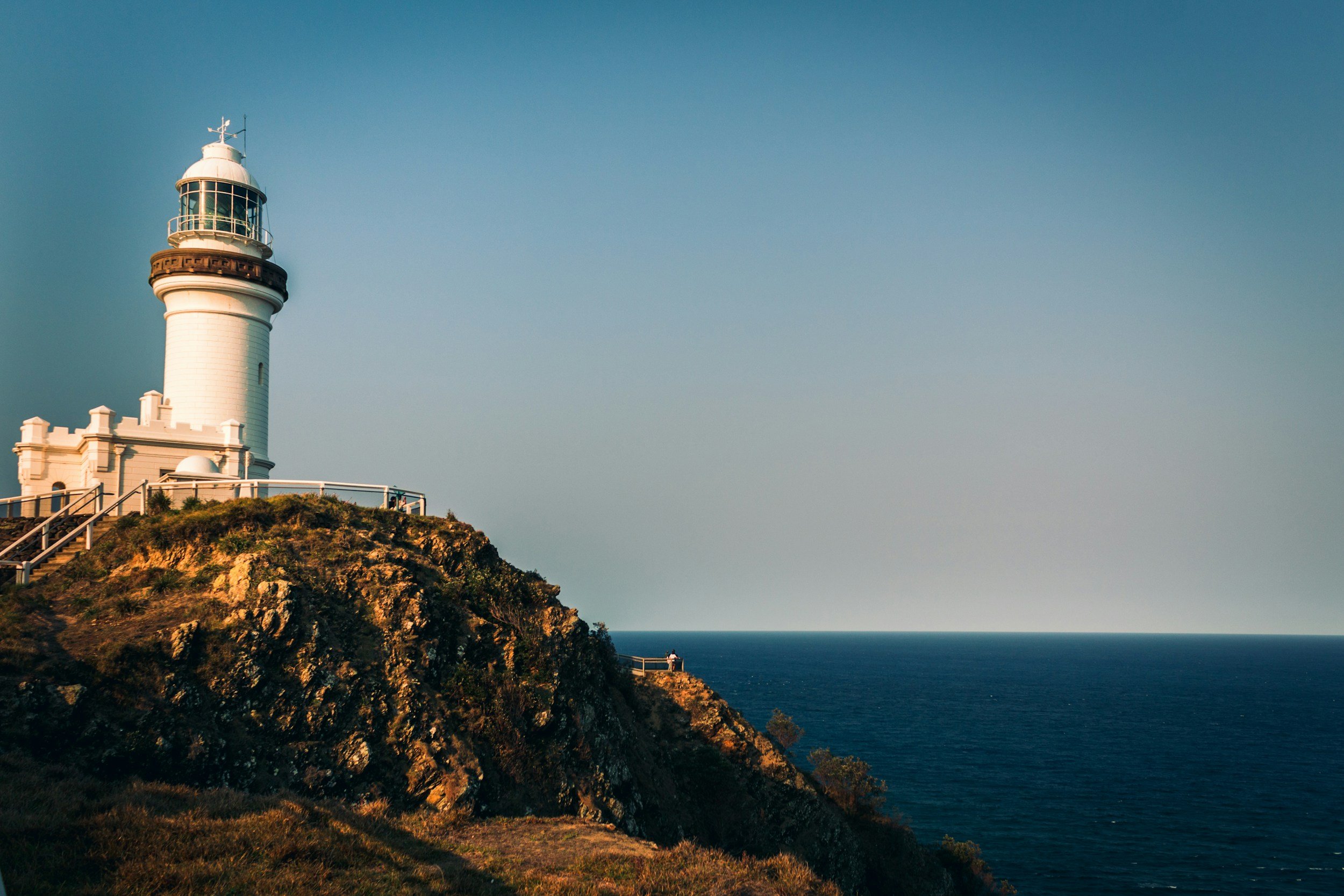 A white lighthouse on a rocky cliff overlooking the ocean with a clear sky in the background.