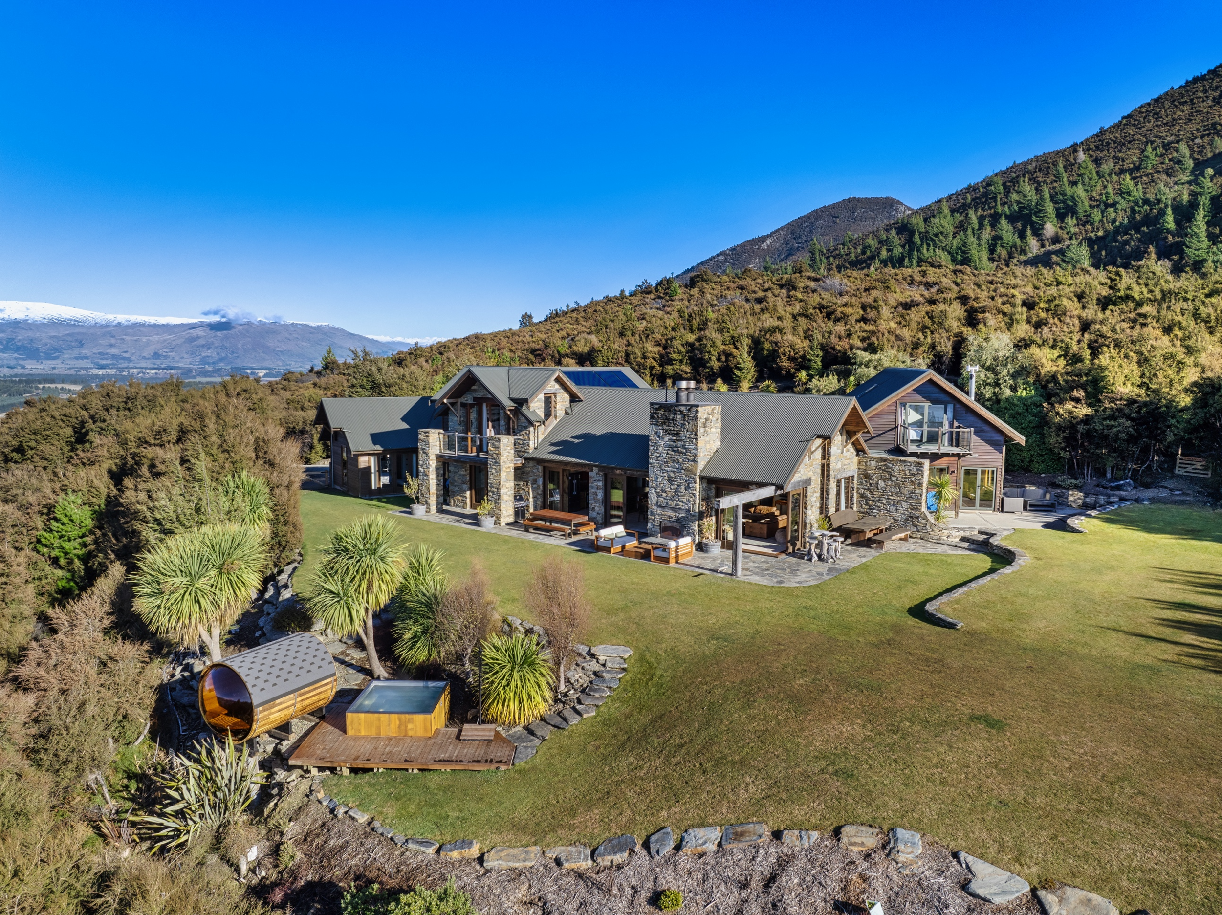 A modern mountain house with stone chimneys and a gray roof in a lush green landscape, surrounded by trees and hills, with a small outdoor hot tub and wooden deck in the yard.