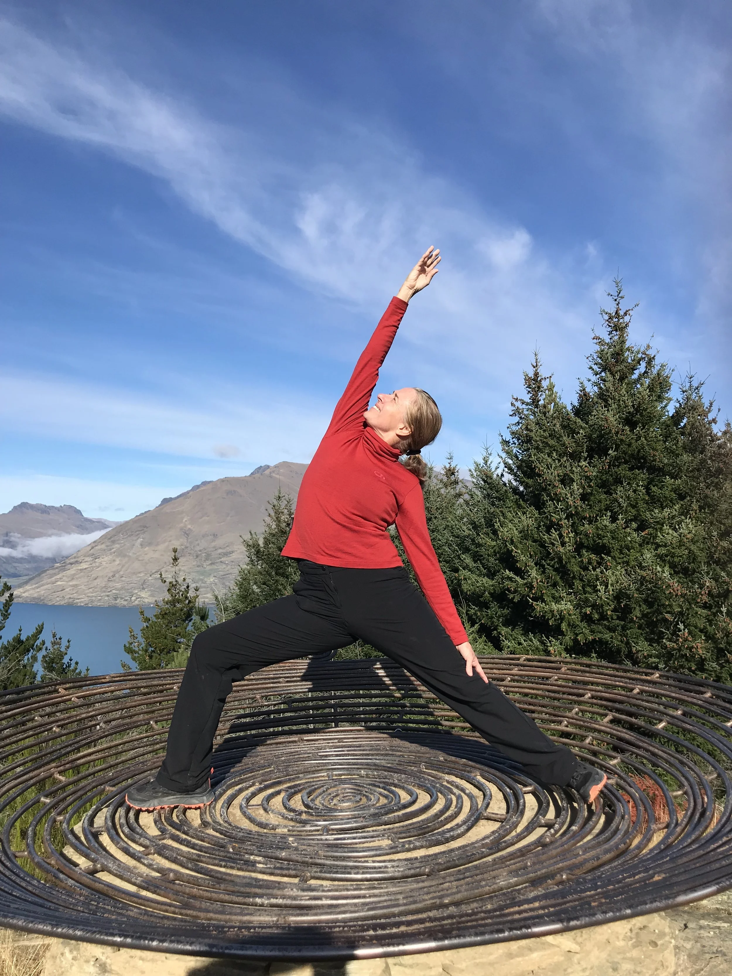 A woman in a red jacket and black pants practicing yoga on a circular outdoor platform with a mountain and lake in the background.