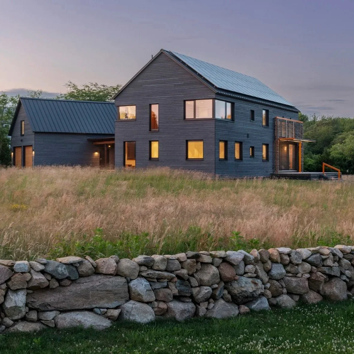 Modern dark-colored house with multiple windows and a metal roof, surrounded by a grassy field and a stone wall, during dusk with illuminated interior lights.
