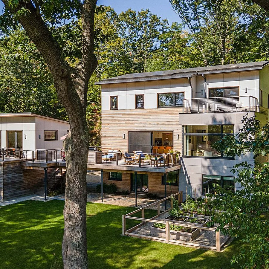 Modern multi-story house with large windows, wooden siding, and a spacious outdoor deck surrounded by greenery and trees.