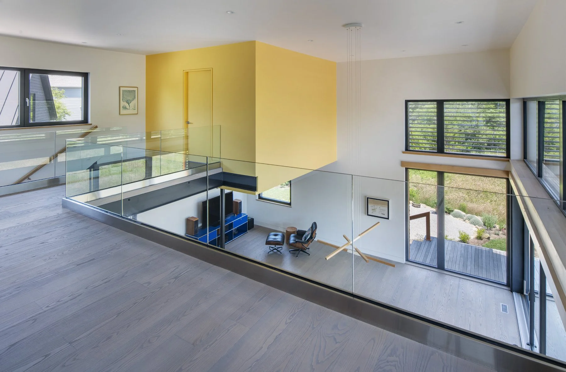 Interior view of a modern house with large windows, glass railing, yellow accent wall, and from a second-floor balcony overlooking a living room area with chairs and a TV.