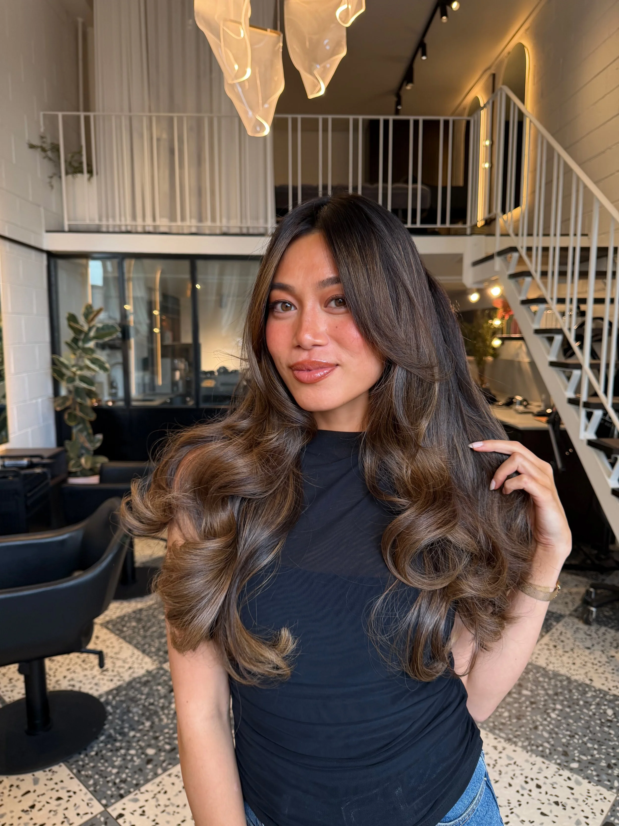 A woman with long, wavy brown hair with highlights, wearing a black top, in a modern salon or studio with a loft, stairs, and large windows.