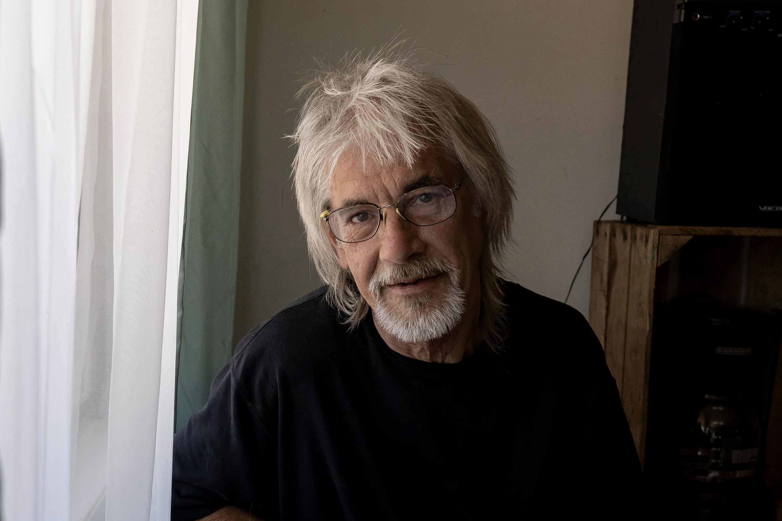 An elderly man with long gray hair, glasses, and a beard wearing a black shirt sitting indoors near a window with light-colored curtains.