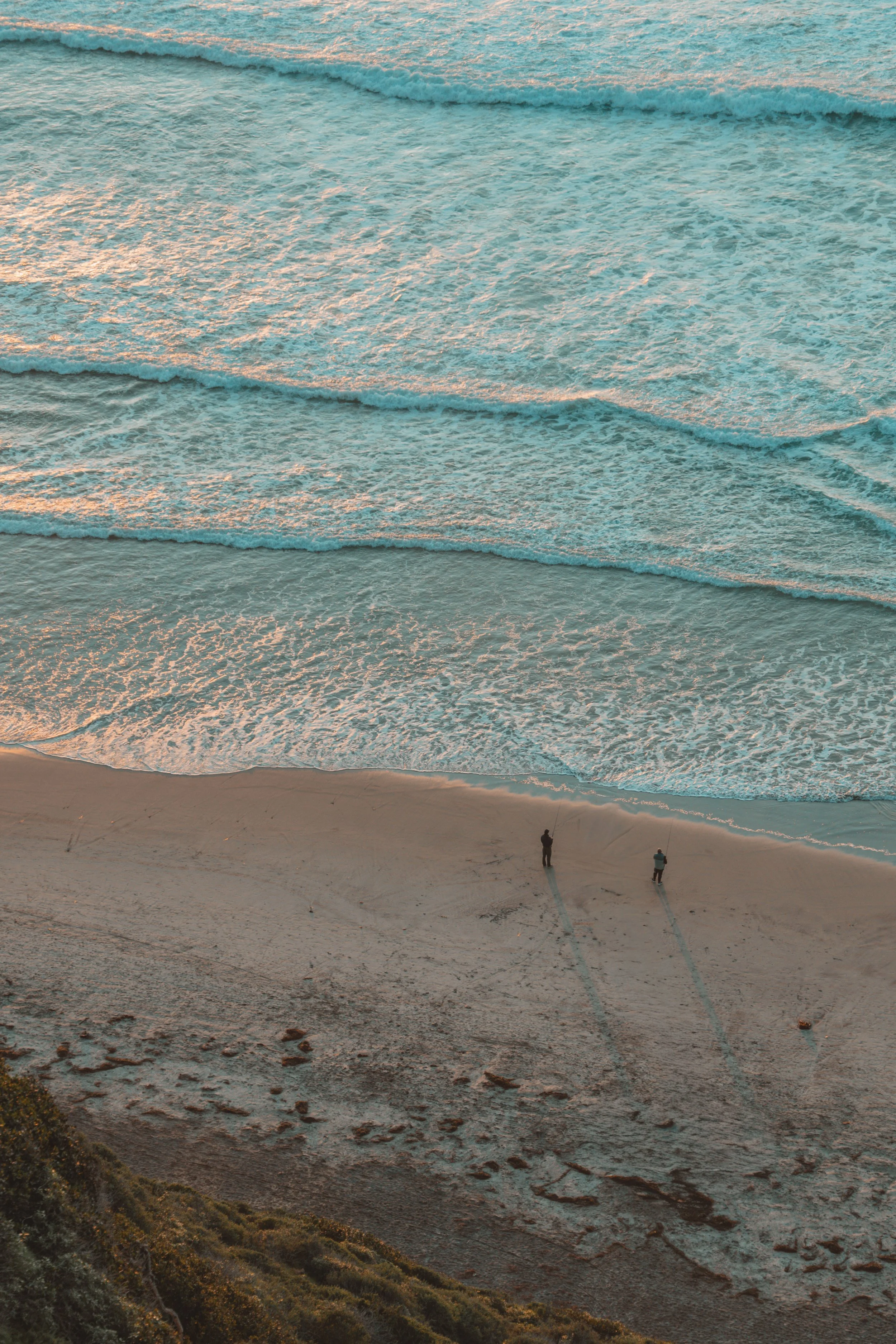 Two people standing on a sandy beach near the shoreline with waves coming in during sunset.
