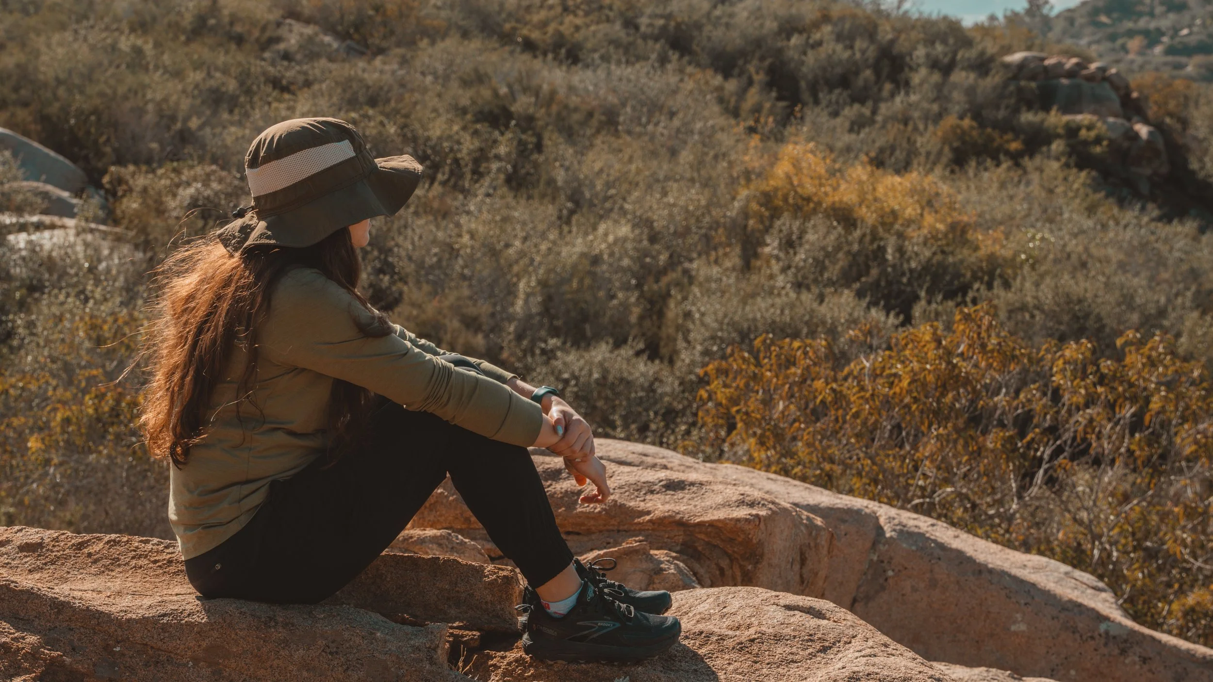 A woman sitting on a rock in a dry, rocky landscape with bushes and hills in the distance, wearing a wide-brimmed hat, olive green jacket, black pants, and hiking shoes, looking away.