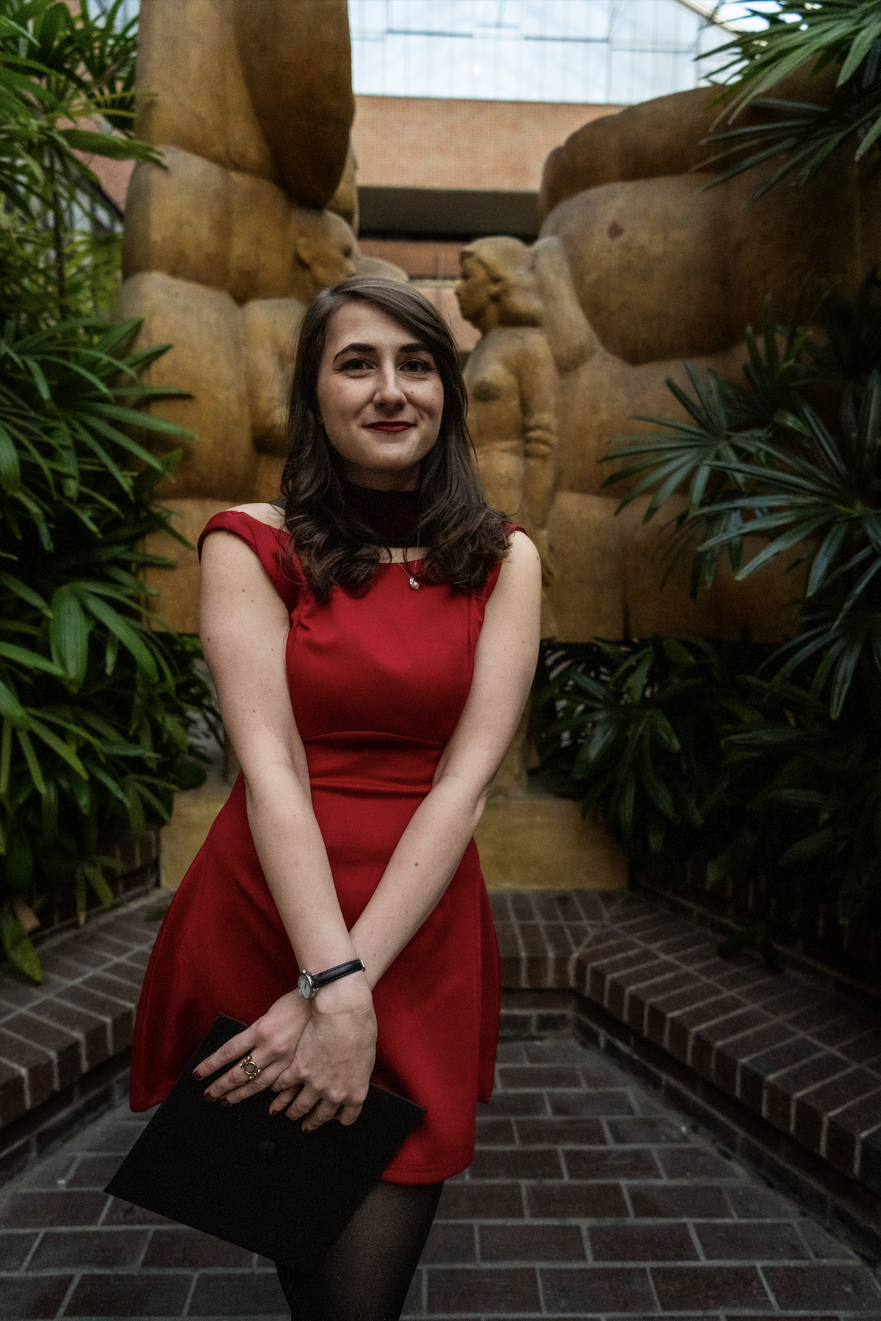 Young woman in a red dress holding a black clutch standing in front of stone sculptures and greenery inside a building.