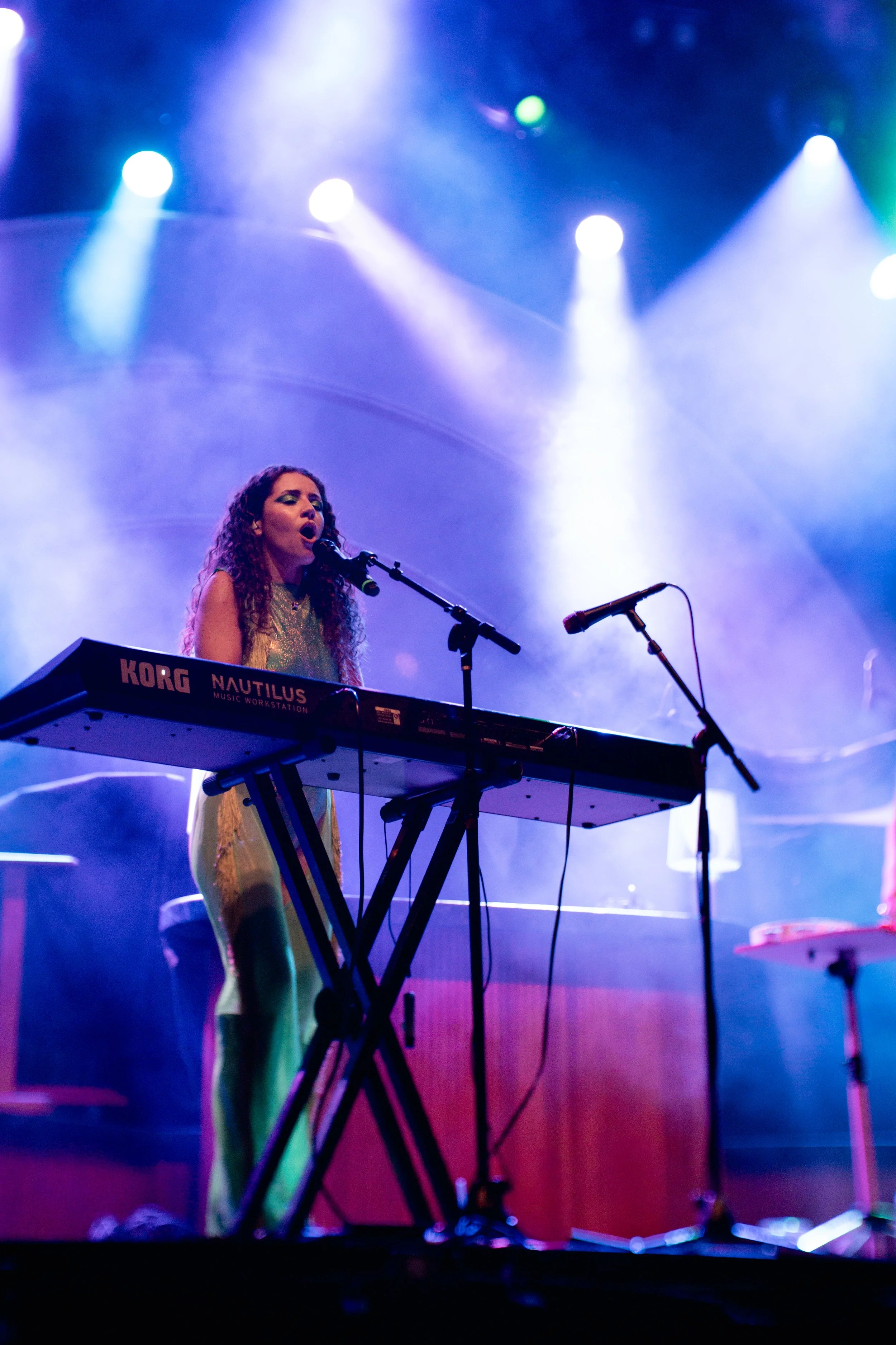 A woman performs on stage with a keyboard, singing into a microphone, illuminated by colorful stage lights.
