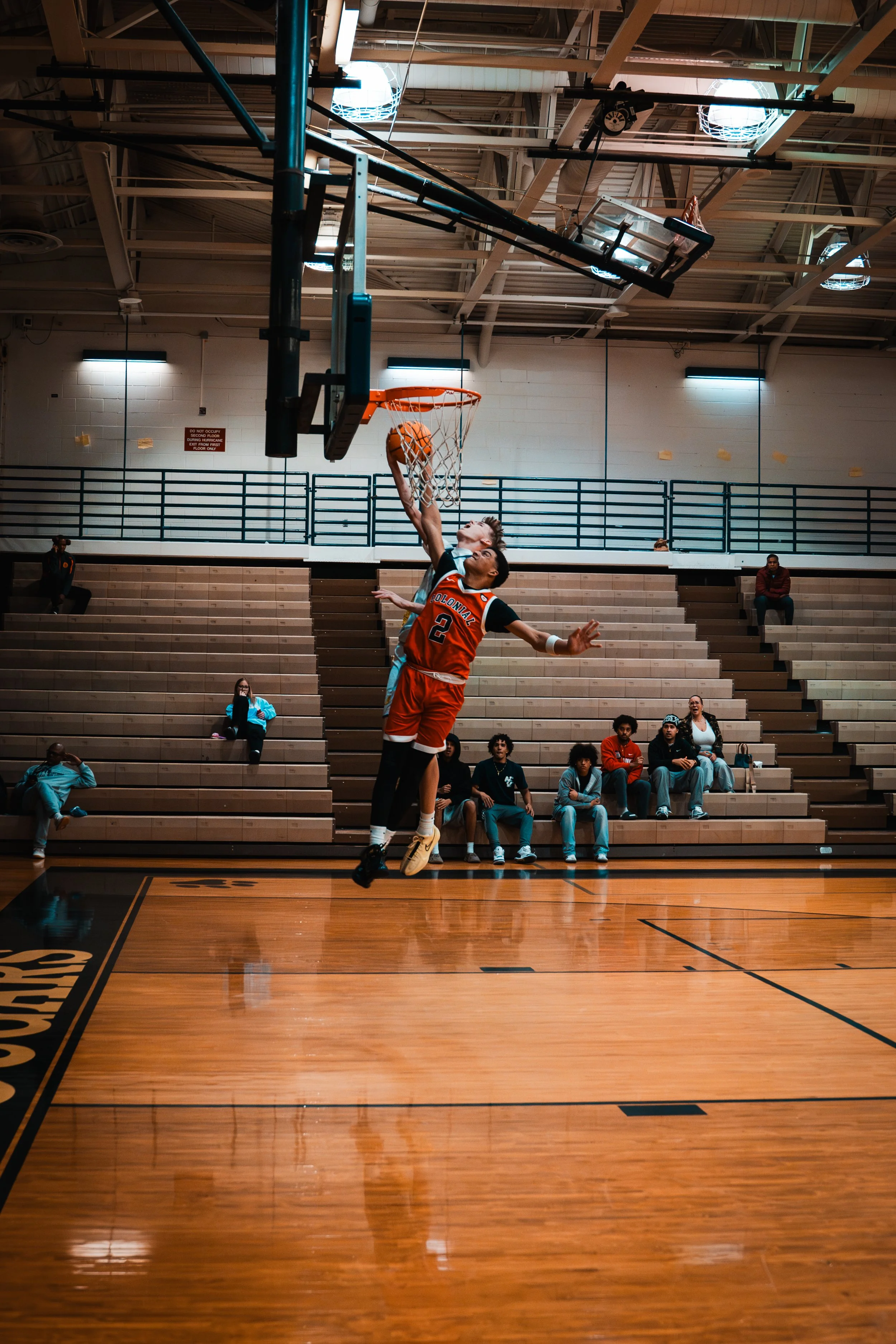 A basketball player in a red uniform jumping to make a shot in an indoor gymnasium, with spectators sitting on bleachers in the background.