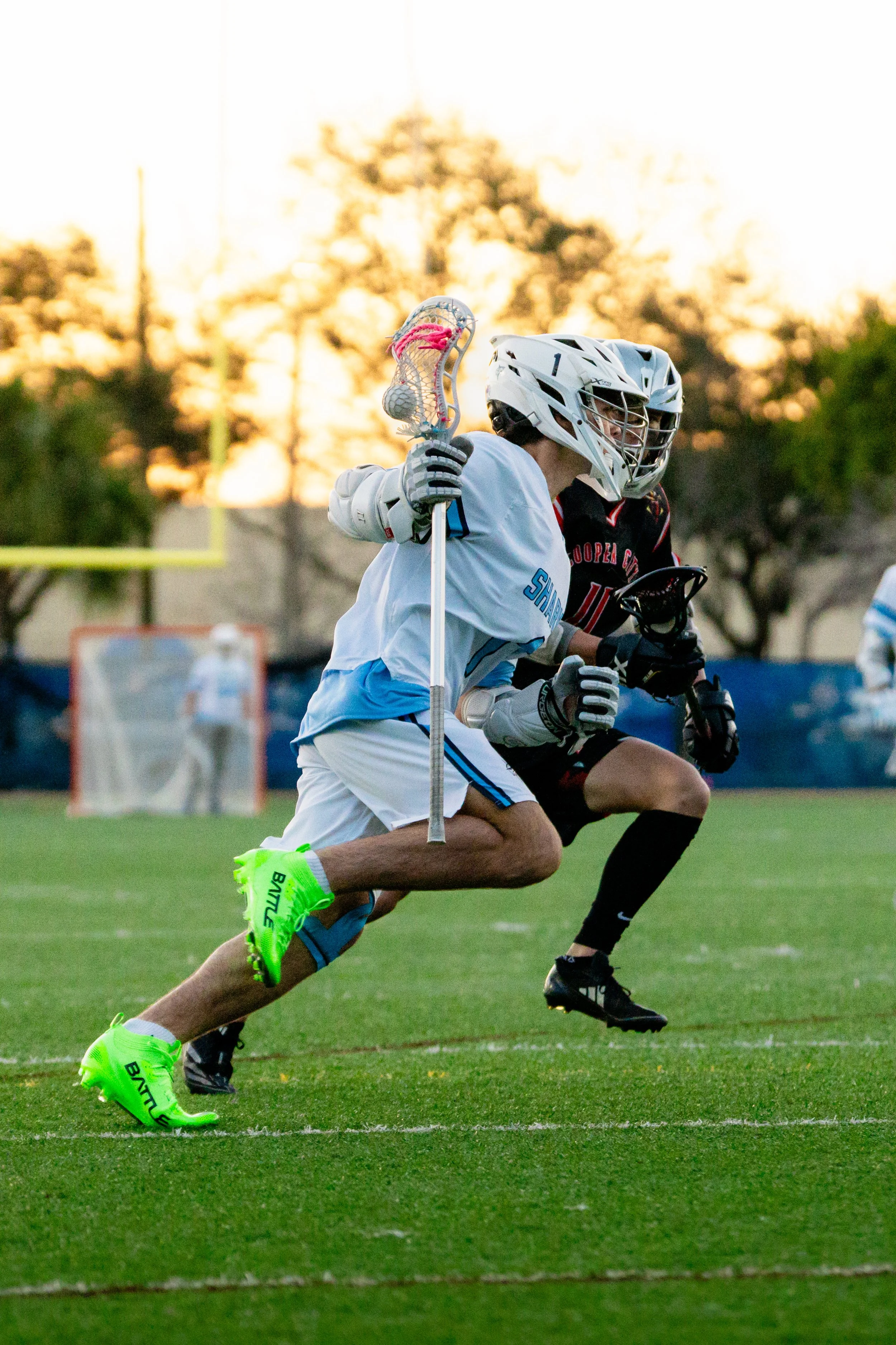 Lacrosse players competing on a field during sunset with trees in the background.
