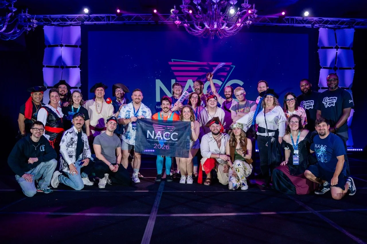 Group of people dressed in costumes and casual wear on stage at NACC 2026 event, some holding a banner with the event logo, under purple lighting and a chandelier.